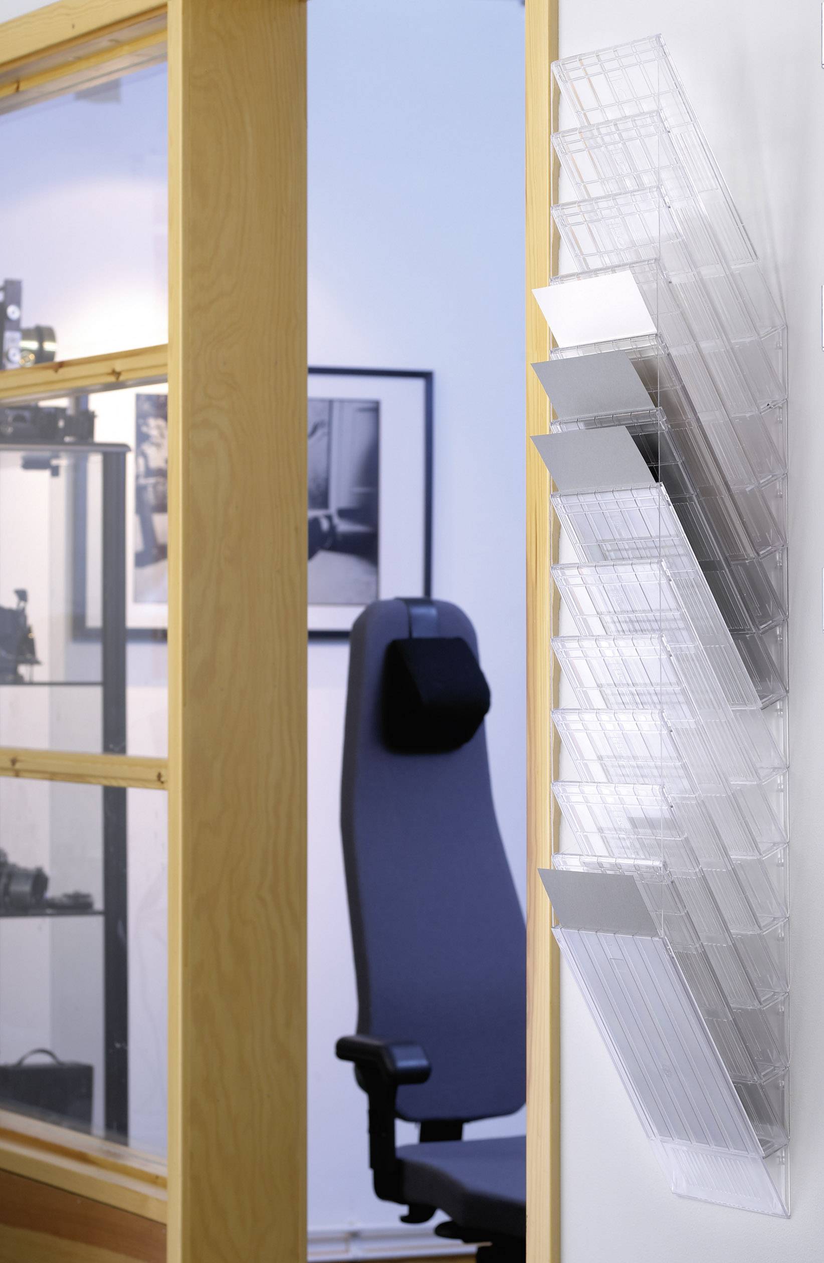 Ein Büro mit einem leeren schwarzen Stuhl vor einer Wand mit transparenten Plastikablagen. Im Hintergrund Regale und ein schwarz-weißes Foto.