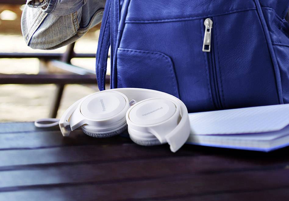 Kopfhörer liegen auf einem Holztisch neben einem blauen Rucksack und einem Notizbuch. Im Hintergrund sind verschwommene Bänke sichtbar.