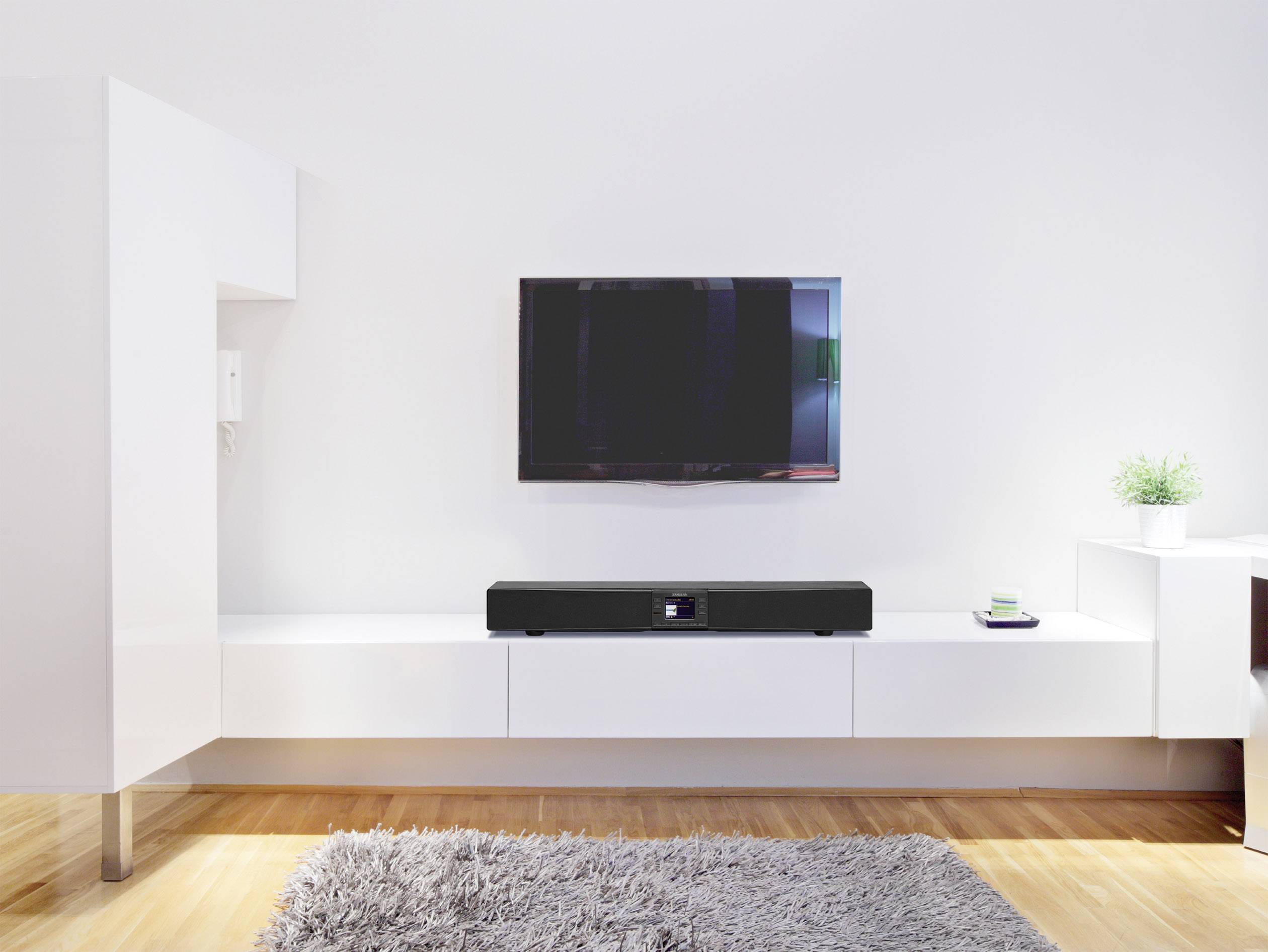 Wohnzimmer mit modernem TV an der Wand; darunter eine Soundbar auf einem weißen Lowboard. Teppich auf Holzboden, minimalistische Einrichtung.