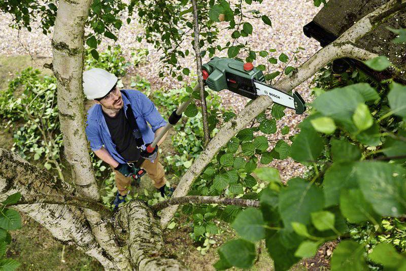 Ein Mann in Arbeitskleidung schneidet einen Baumast mit einer elektrischen Kettensäge. Er trägt einen Schutzhelm und steht auf dem Boden.