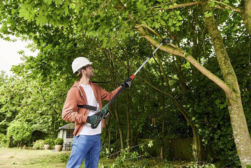 Ein Mann mit Schutzbrille und Helm beschneidet Äste mit einem elektrischen Astschneider in einem Garten.