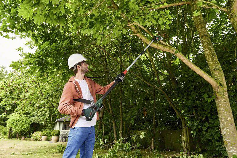 Ein Mann mit Helm und Schutzbrille verwendet eine Kettensäge an einem Stab, um Äste eines Baumes in einem Garten zu schneiden.