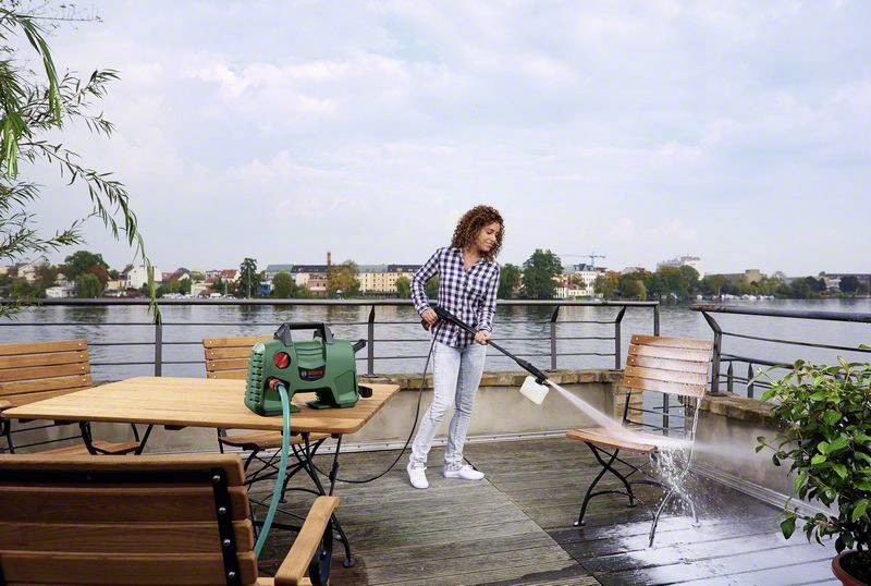 Eine Frau säubert mit einem Hochdruckreiniger eine Terrasse mit Holzmöbeln. Im Hintergrund ist ein Fluss mit Stadtansicht zu sehen.