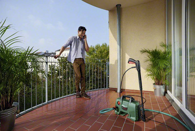 Ein Mann telefoniert auf einem Balkon. Neben ihm steht ein Hochdruckreiniger mit Schlauch. Im Hintergrund sind Pflanzen und Gebäude sichtbar.