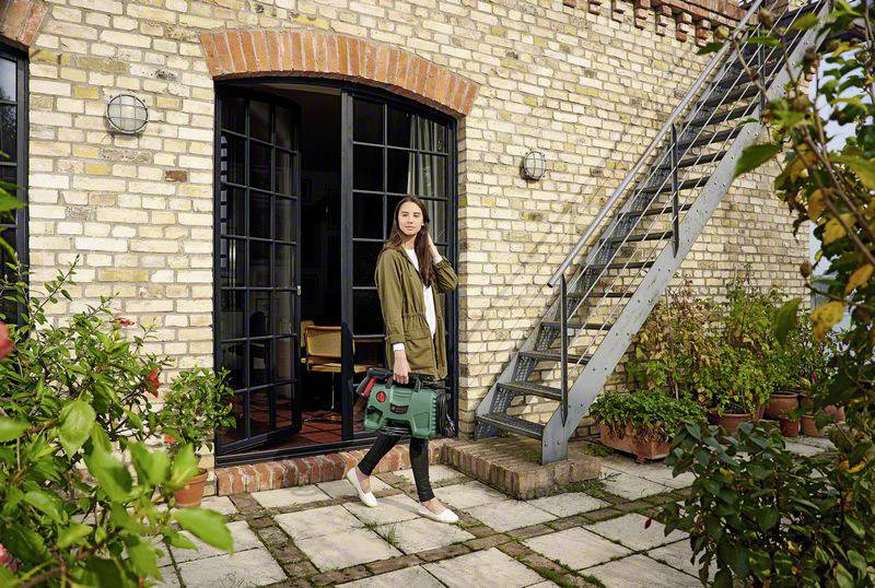 Frau in legerer Kleidung trägt eine grüne Werkzeugtasche und steht auf einer Terrasse vor einem Haus aus Backstein mit einer Metalltreppe.