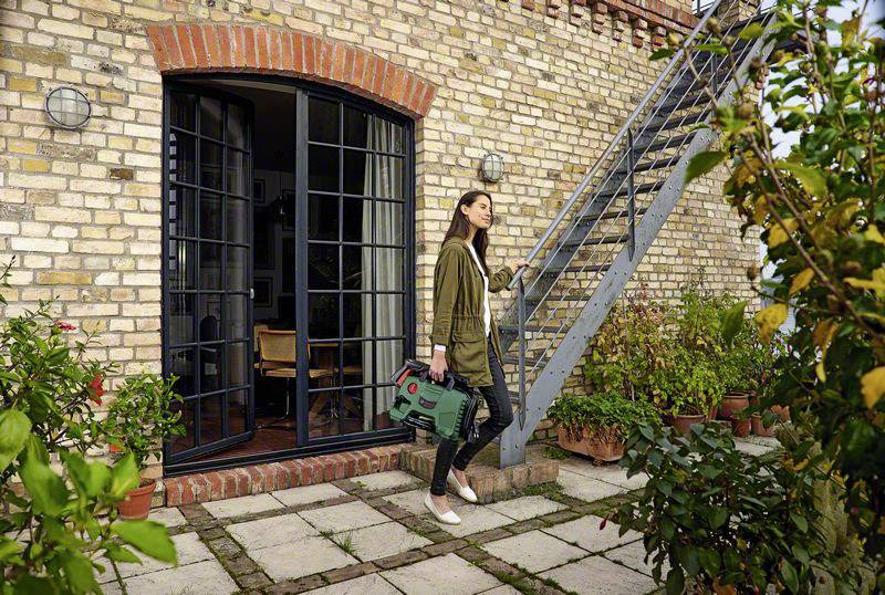 Frau trägt eine grüne Gießkanne und geht auf einem gepflasterten Weg neben einem Backsteinhaus mit einem metallenen Treppengeländer.