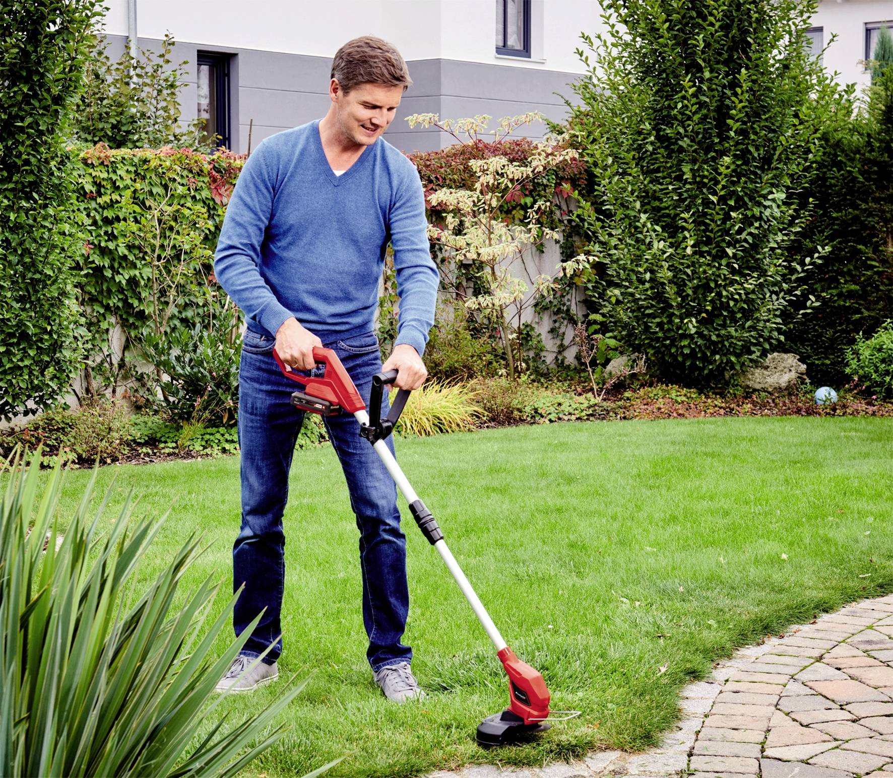 Ein Mann trimmt mit einem elektrischen Rasentrimmer den Rasen in einem Garten. Im Hintergrund sind Büsche und ein Haus zu sehen.