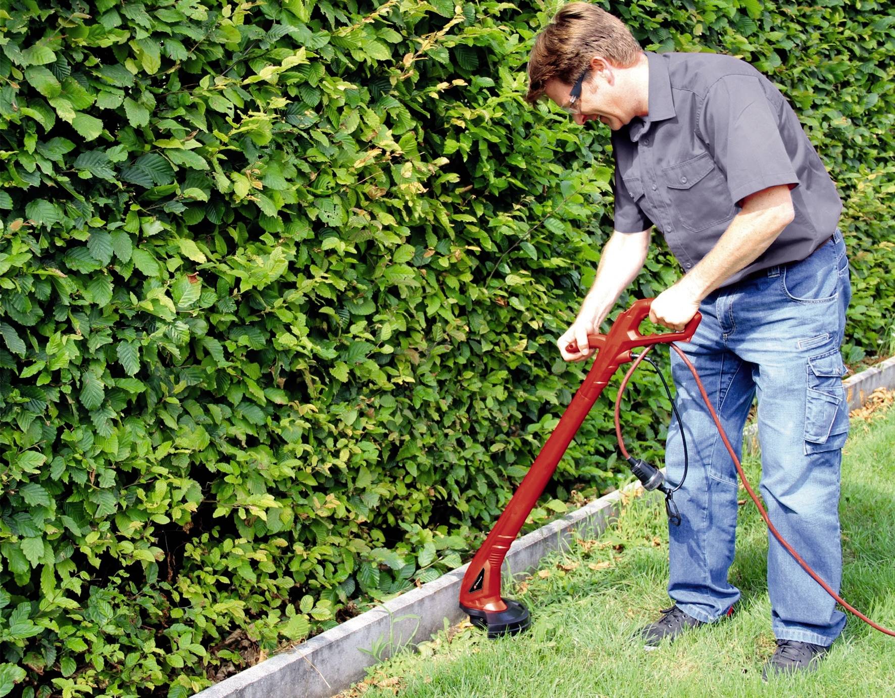Eine Person trimmt mit einem Elektroschneider das Gras entlang einer Hecke in einem Garten.