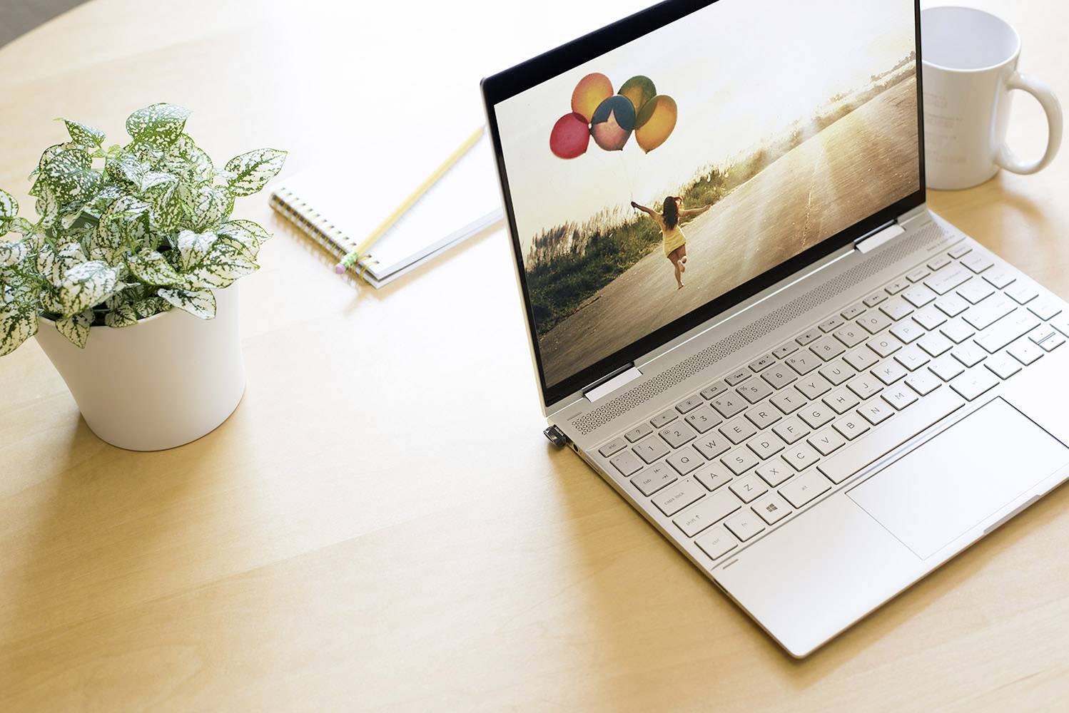 Silberner Laptop auf Holztisch mit Bild von Kind, das Luftballons hält. Daneben Kaffeetasse, Pflanze und Notizblock.