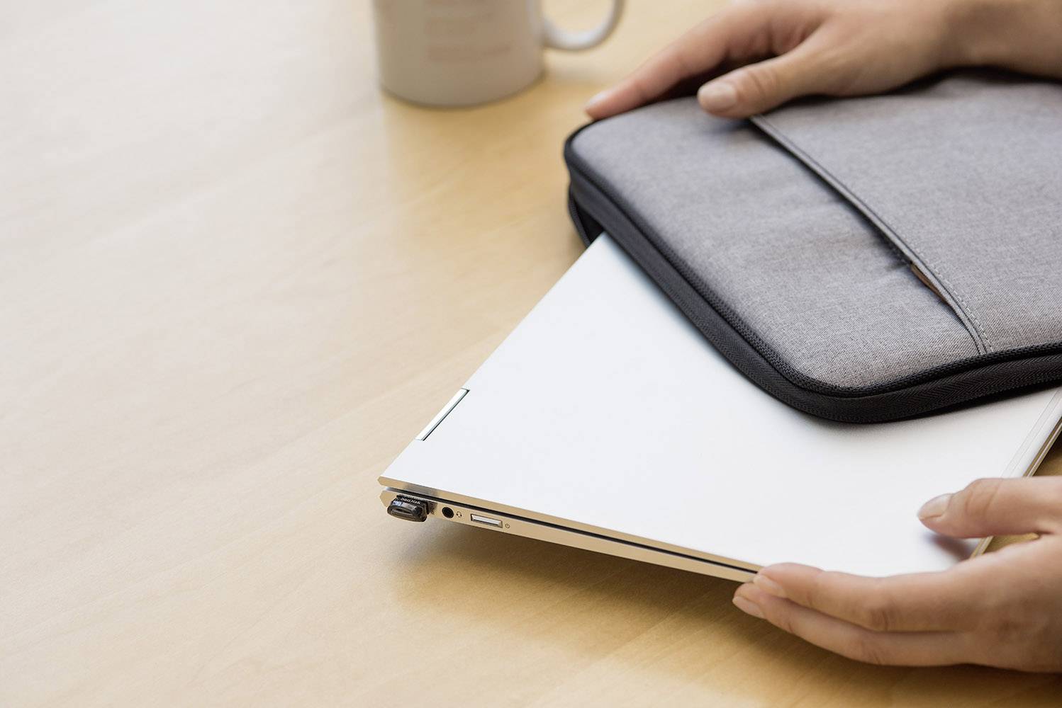 Person legt einen geschlossenen Laptop in eine graue Hülle auf einem Holztisch. Im Hintergrund steht eine Tasse.