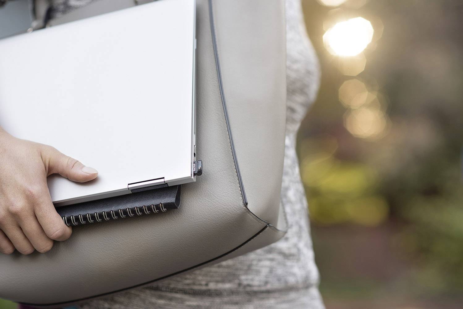 Eine Person trägt eine hellgraue Tasche mit einem Laptop und einem Notizbuch, im Freien bei Sonnenuntergang.