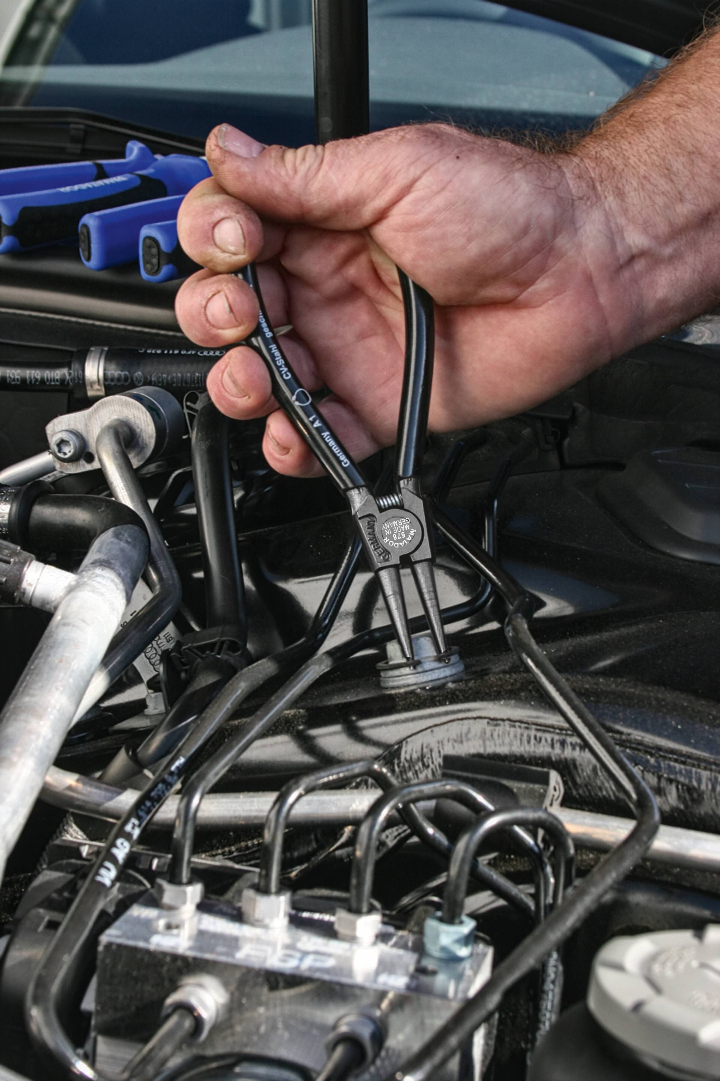 Eine Hand hält einen Abzieher, um einen Klemmverbinder im Motorraum eines Autos zu lösen. Mechanikerwerkzeuge und Kabel sind sichtbar.