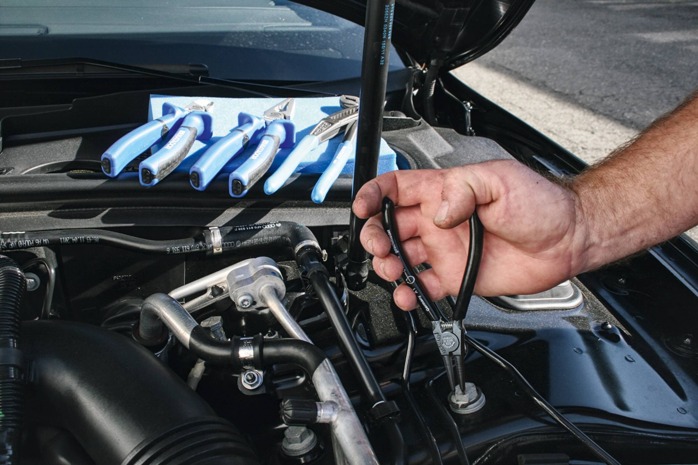 Eine Hand benutzt eine Zange, um an einem Autoteil unter der Motorhaube zu arbeiten. Ein Set von Handwerkzeugen liegt daneben.