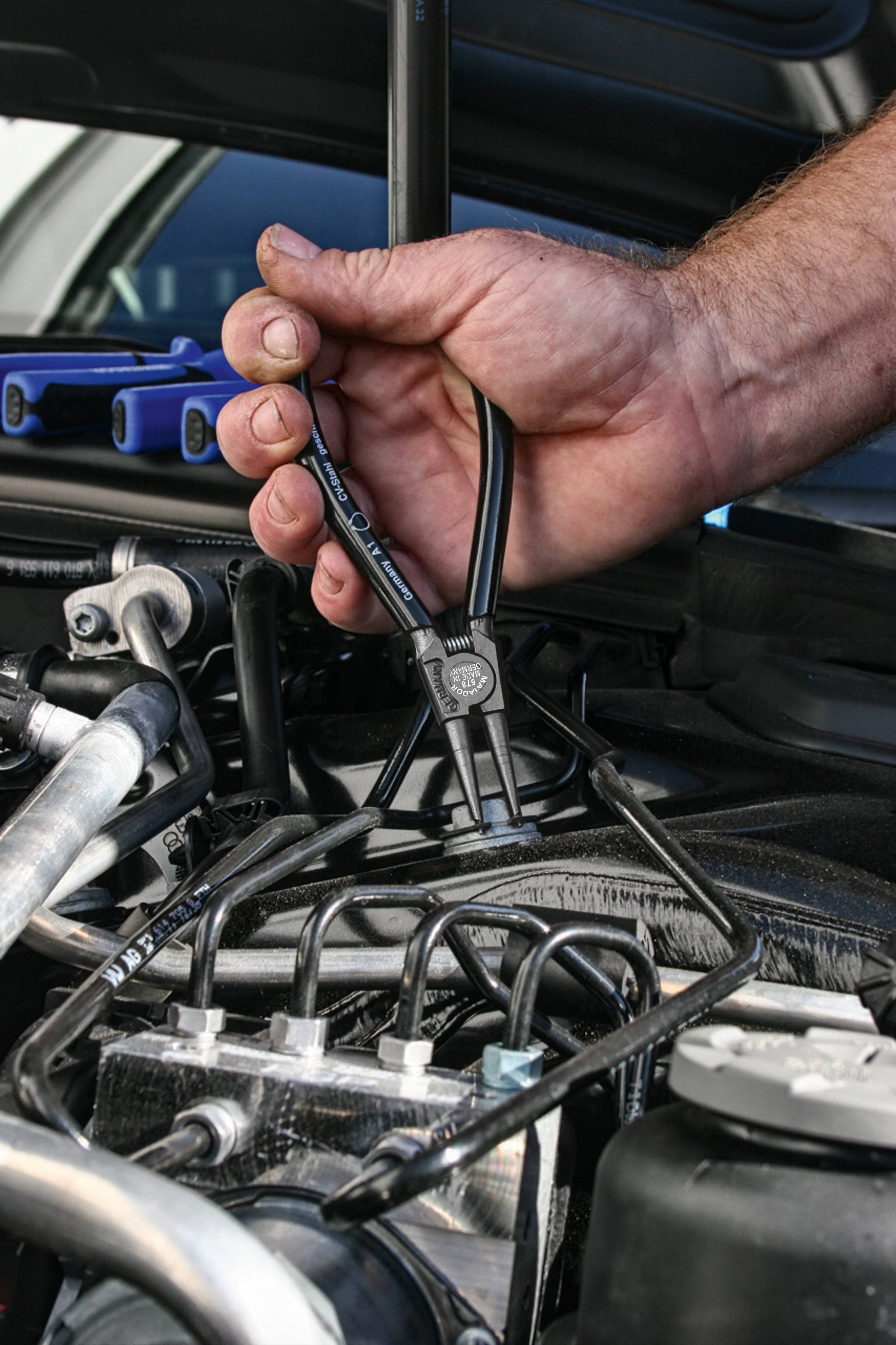 Ein Mechaniker benutzt eine Zange, um an einem Fahrzeugmotor zu arbeiten, konzentriert auf Leitungen und technischen Details im Motorraum.