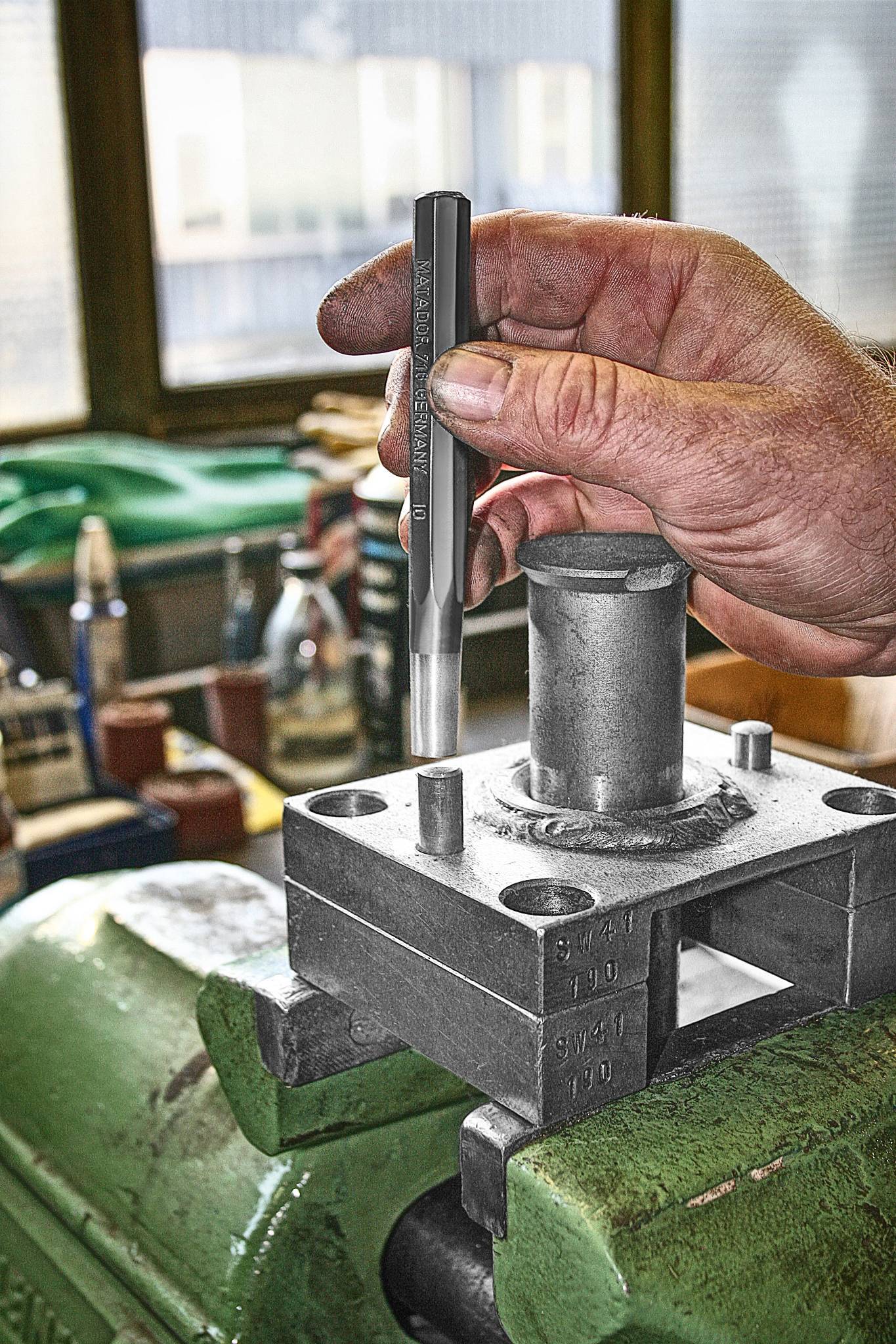Eine Hand hält ein Werkzeug und bearbeitet ein Metallstück in einer Werkstatt. Der Fokus liegt auf Präzisionsarbeit und Handwerkstechnik.