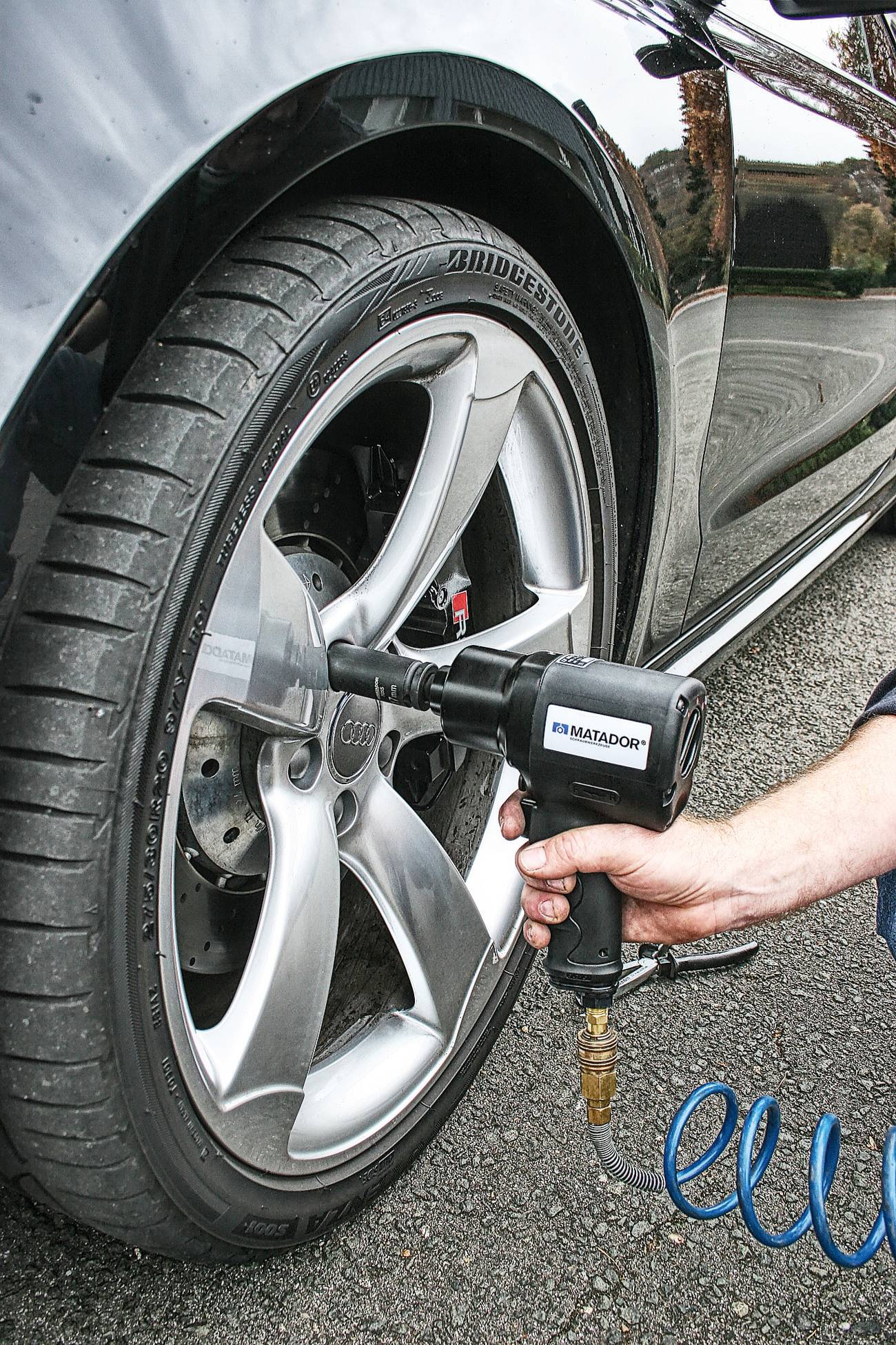 Eine Person wechselt mit einem elektrischen Schlagschrauber das Rad eines Autos. Das Fahrzeug steht auf Asphalt.