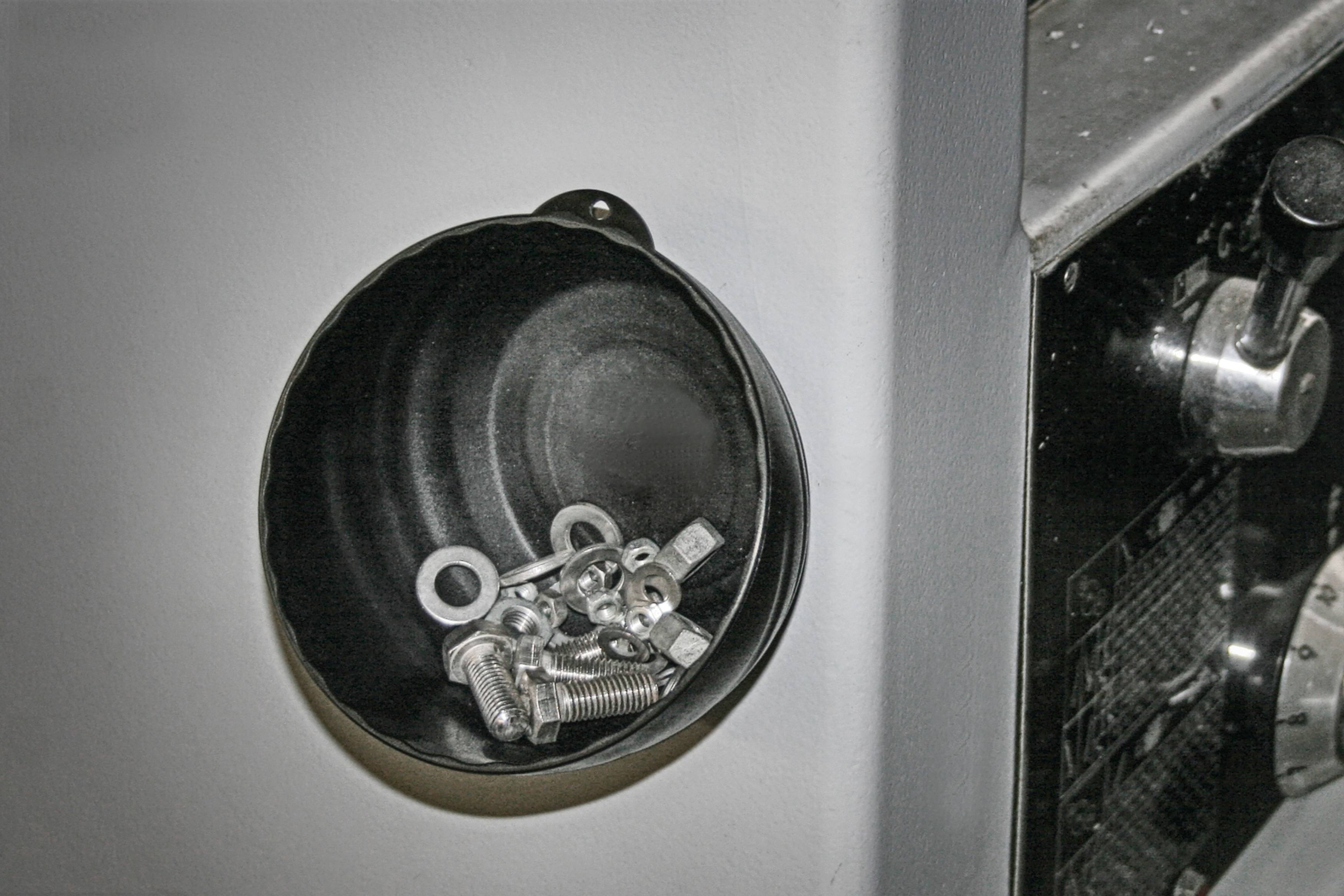 Ein schwarzer Behälter ist an einer Maschinenwand befestigt. Darin liegen Schrauben, Muttern und Unterlegscheiben, die zur Wartung verwendet werden.