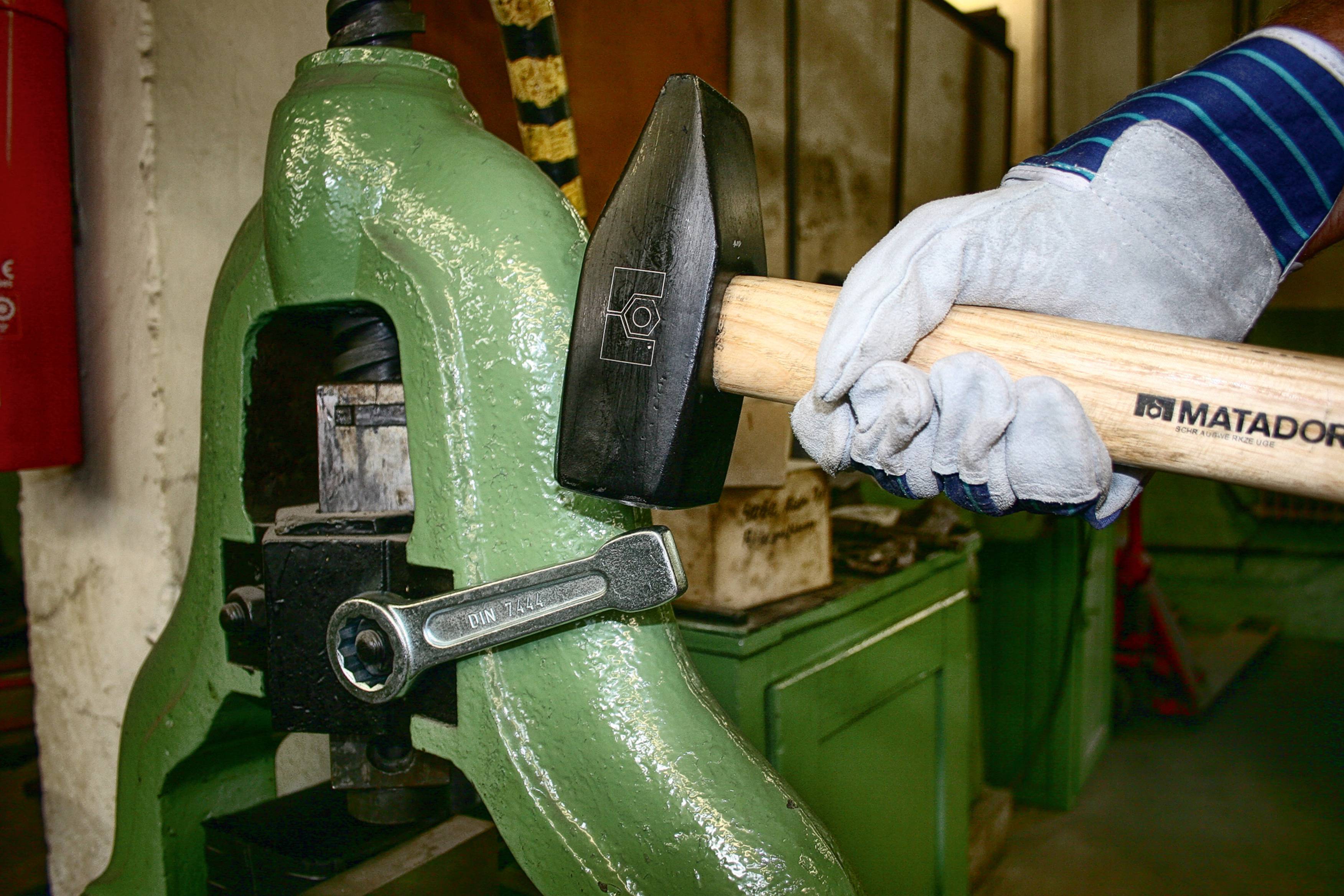 Eine Person schlägt mit einem Hammer auf eine Schraubzwinge, die an einem grünen Metallgerät in einer Werkstatt befestigt ist.