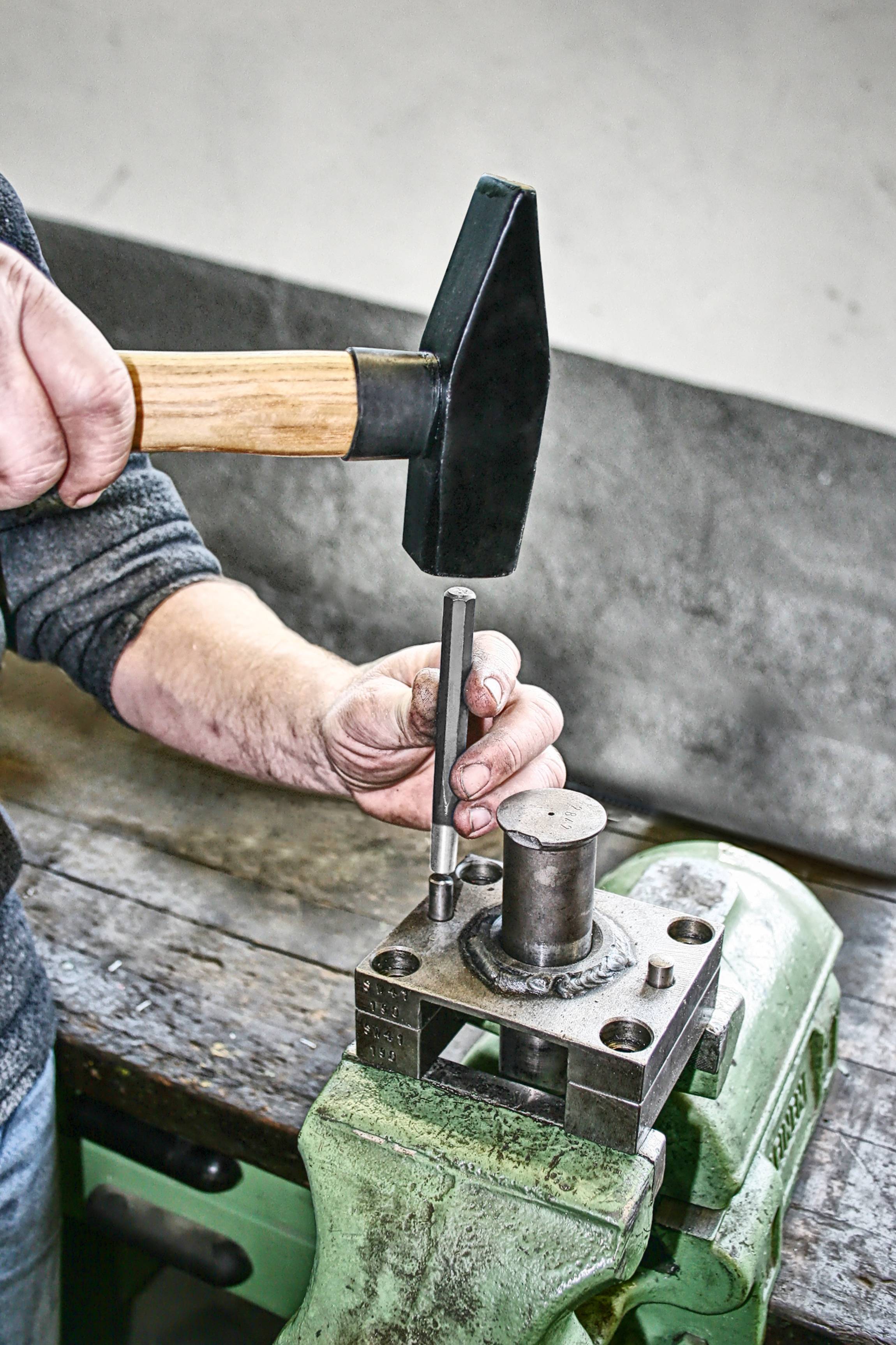 Eine Hand hält einen Hammer, um einen Metallstift in ein Werkstück zu treiben, das in einem Schraubstock auf einer Werkbank fixiert ist.