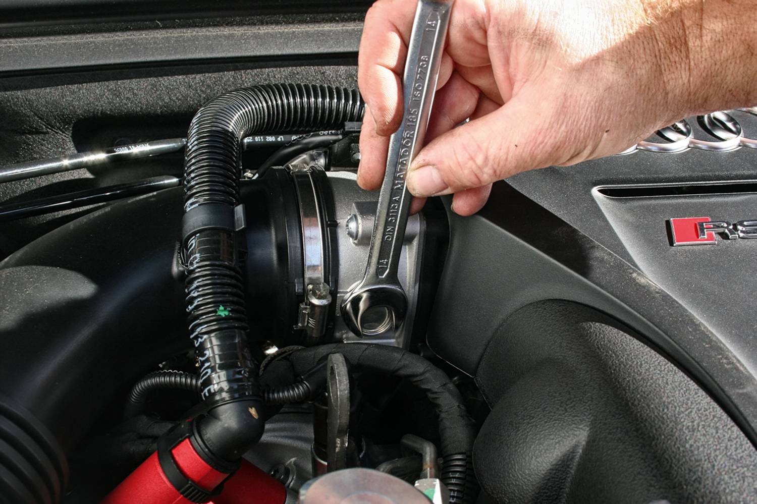 Eine Hand hält einen Schraubenschlüssel, der eine Schraube im Motorraum eines Autos festzieht. Der Motor ist teilweise sichtbar.