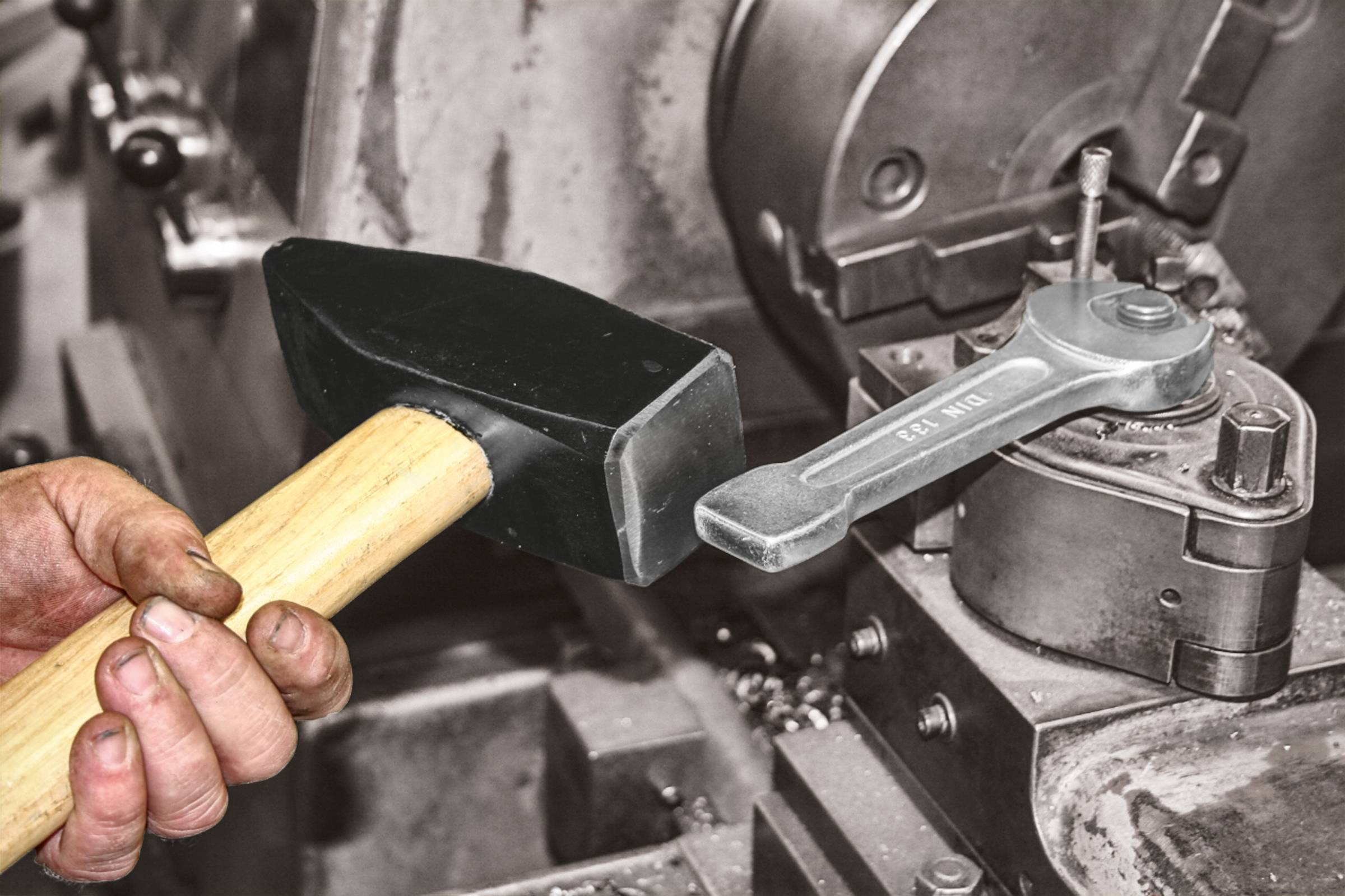 Eine Hand hält einen Hammer vor einer großen Metallmaschine, die in einer Werkstatt zu sehen ist.