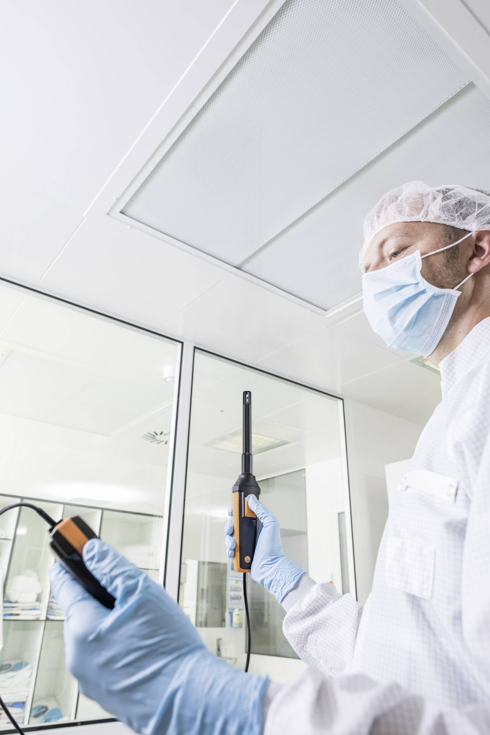 Ein Labortechniker in steriler Kleidung misst mit einem Gerät die Luftqualität in einem Reinraum. Hintergrund zeigt Laborumgebung.