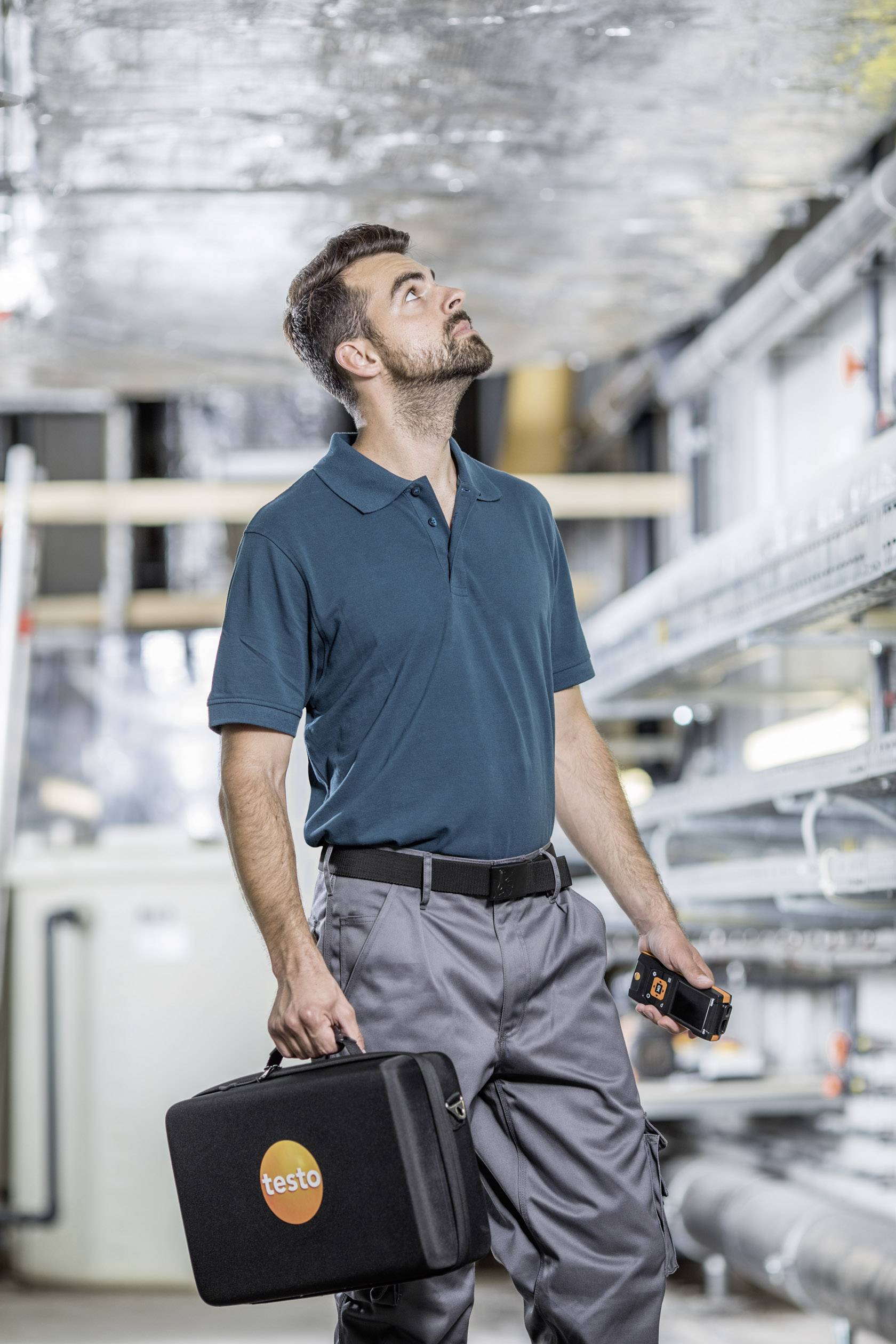 Techniker inspiziert eine industrielle Anlage, trägt ein Poloshirt und hält eine Werkzeugtasche und ein Messgerät.