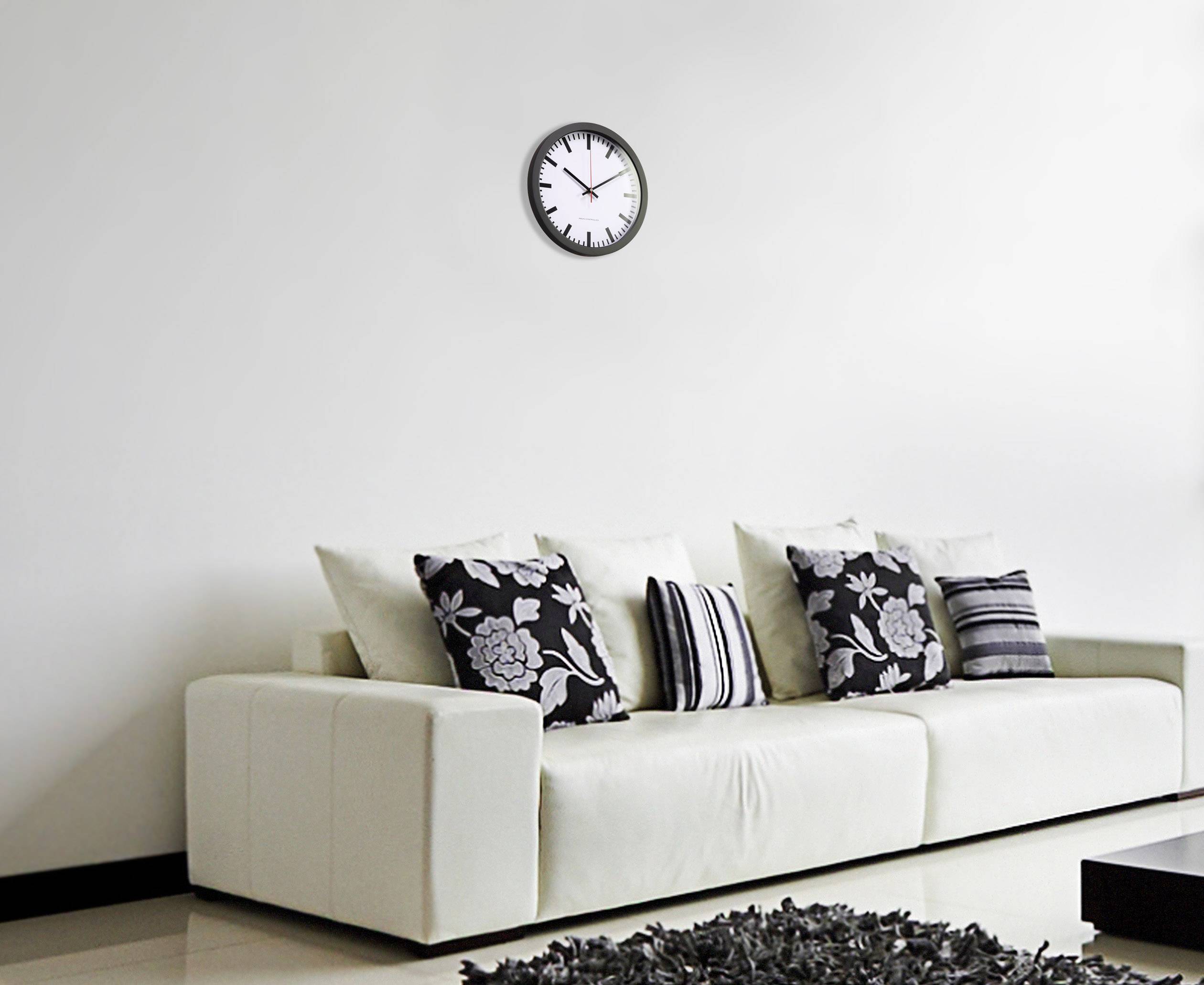 Ein weißes Sofa mit schwarzen und weißen gemusterten Kissen steht vor einer weißen Wand. An der Wand hängt eine analoge Uhr.