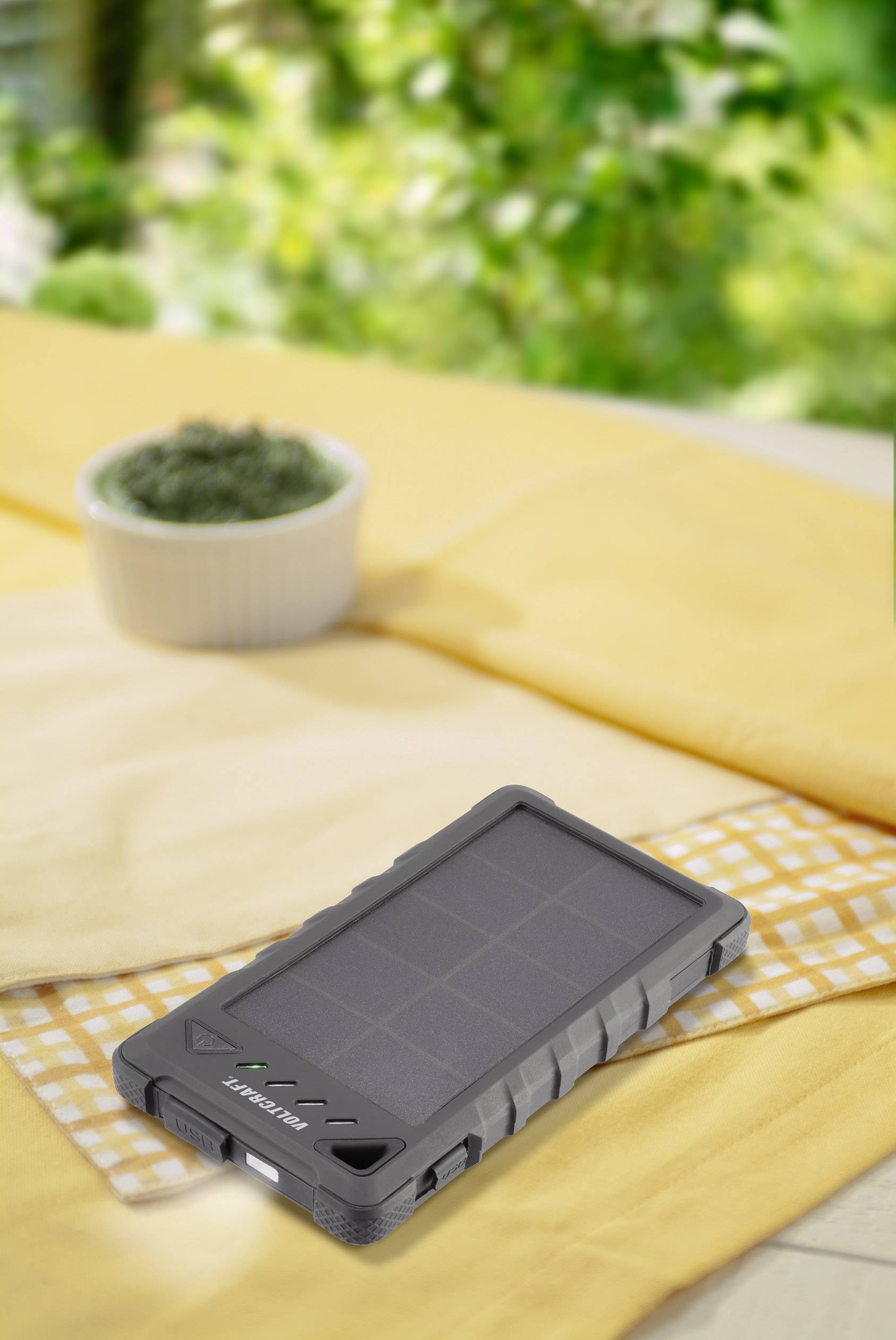 Ein robustes, graues Solarladegerät liegt auf einem Tisch mit gelbem Tuch. Im Hintergrund ist unscharfes Grün zu erkennen.