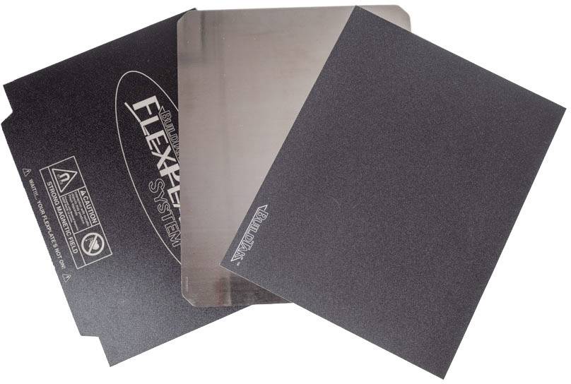 Drei schwarze 3D-Drucker-Bauplatten, eine transparent, teils mit Marken- und Warnhinweisen bedruckt, auf weißem Hintergrund.