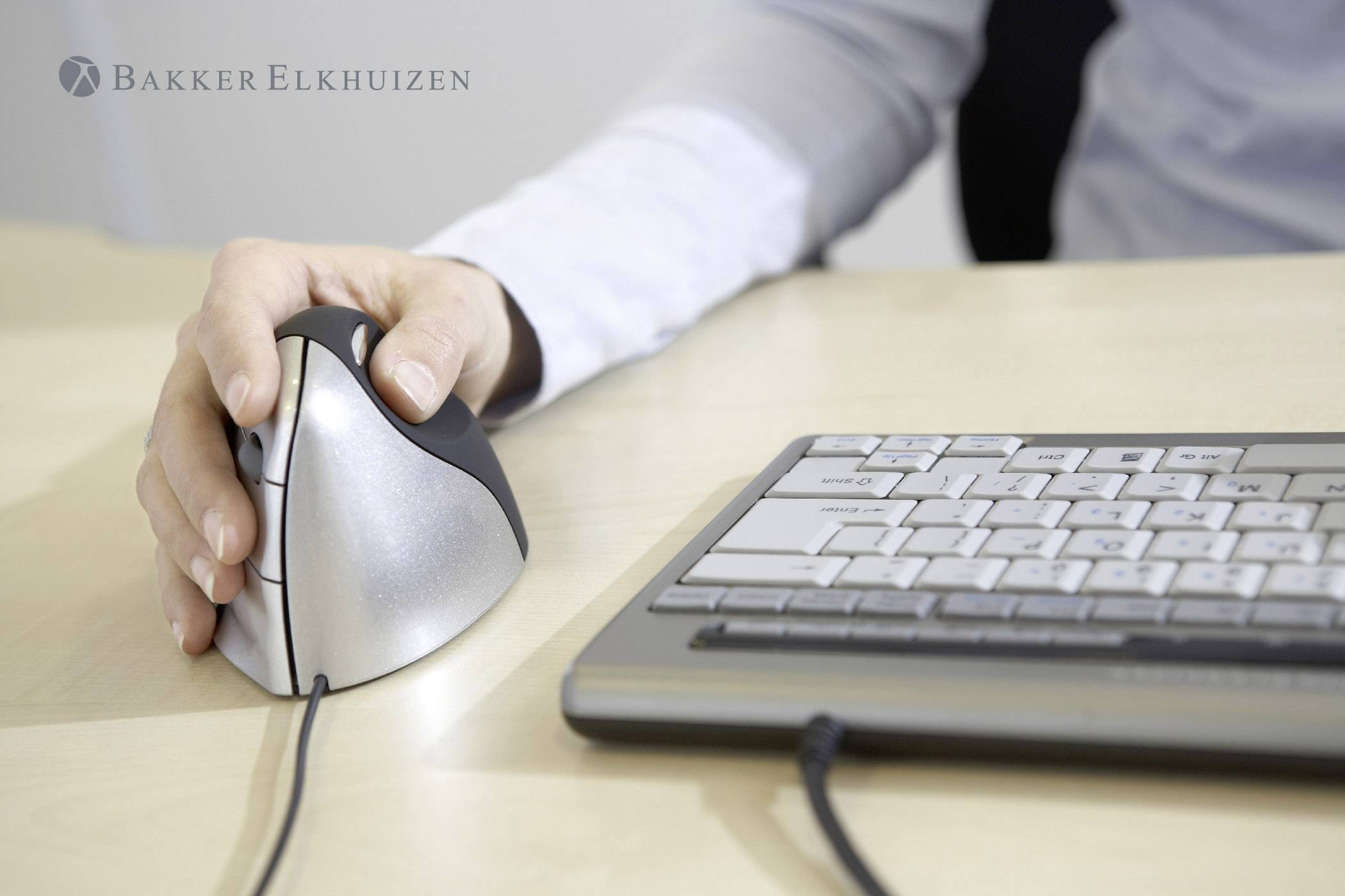 Eine Person benutzt eine ergonomische, vertikale Computermaus neben einer Tastatur auf einem Tisch.