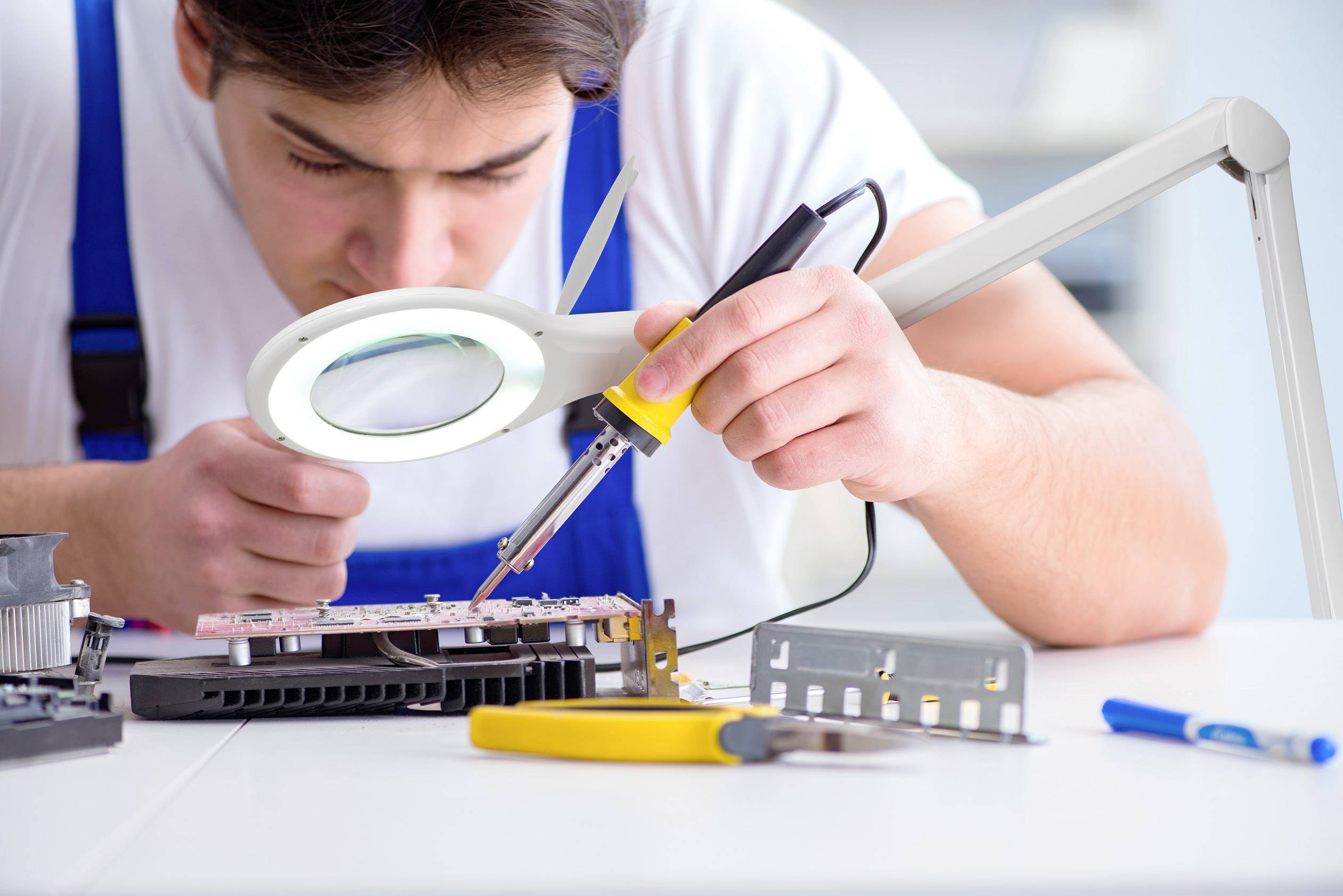 Ein Mann lötet konzentriert eine elektronische Platine unter einer Vergrößerungslampe. Daneben liegen Werkzeuge auf einem Tisch.