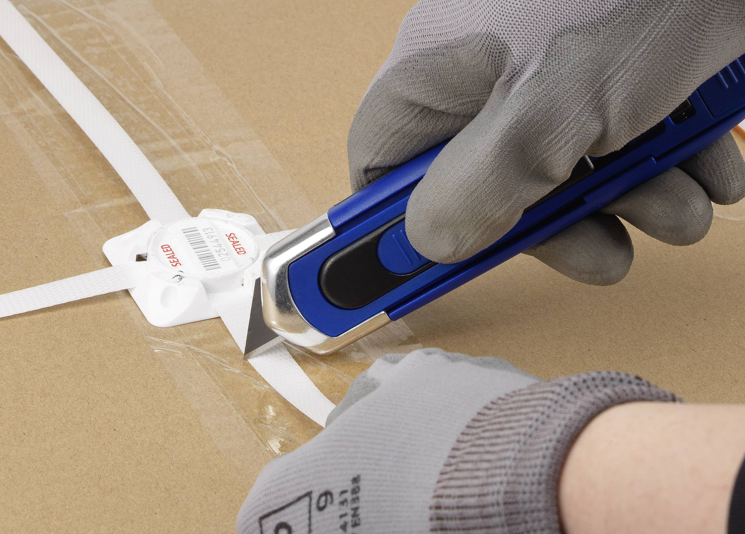 Eine Person schneidet mit einem blauen Cuttermesser ein weißes Plastikband auf einem Karton. Sie trägt graue Schutzhandschuhe.