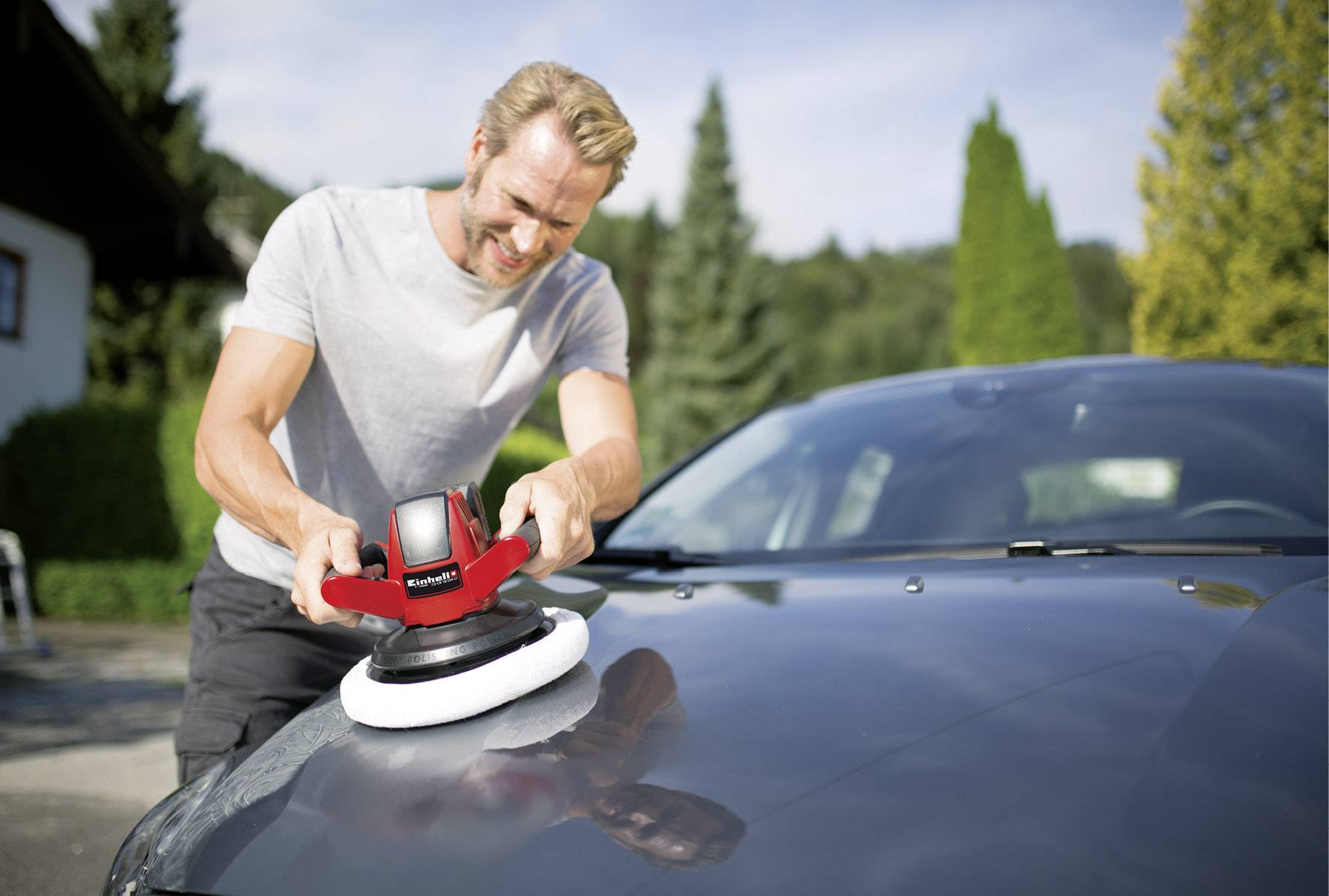Ein Mann poliert die Motorhaube eines Autos mit einer Poliermaschine im Freien. Im Hintergrund sind Bäume und ein Gebäude zu sehen.