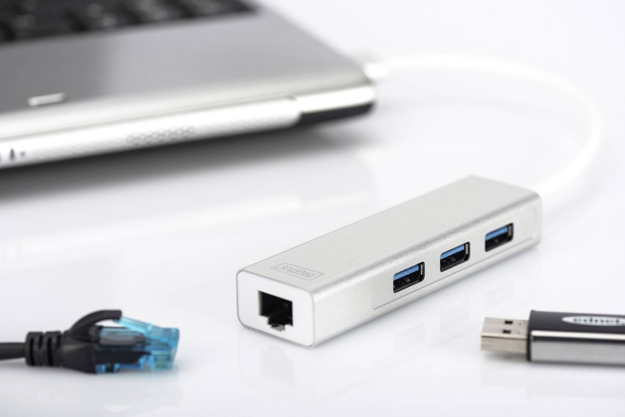Ein silberner USB-Hub mit drei USB-Ports und einem Ethernet-Anschluss liegt neben einem Laptop. Ein USB-Stick und ein Ethernet-Kabel sind daneben platziert.