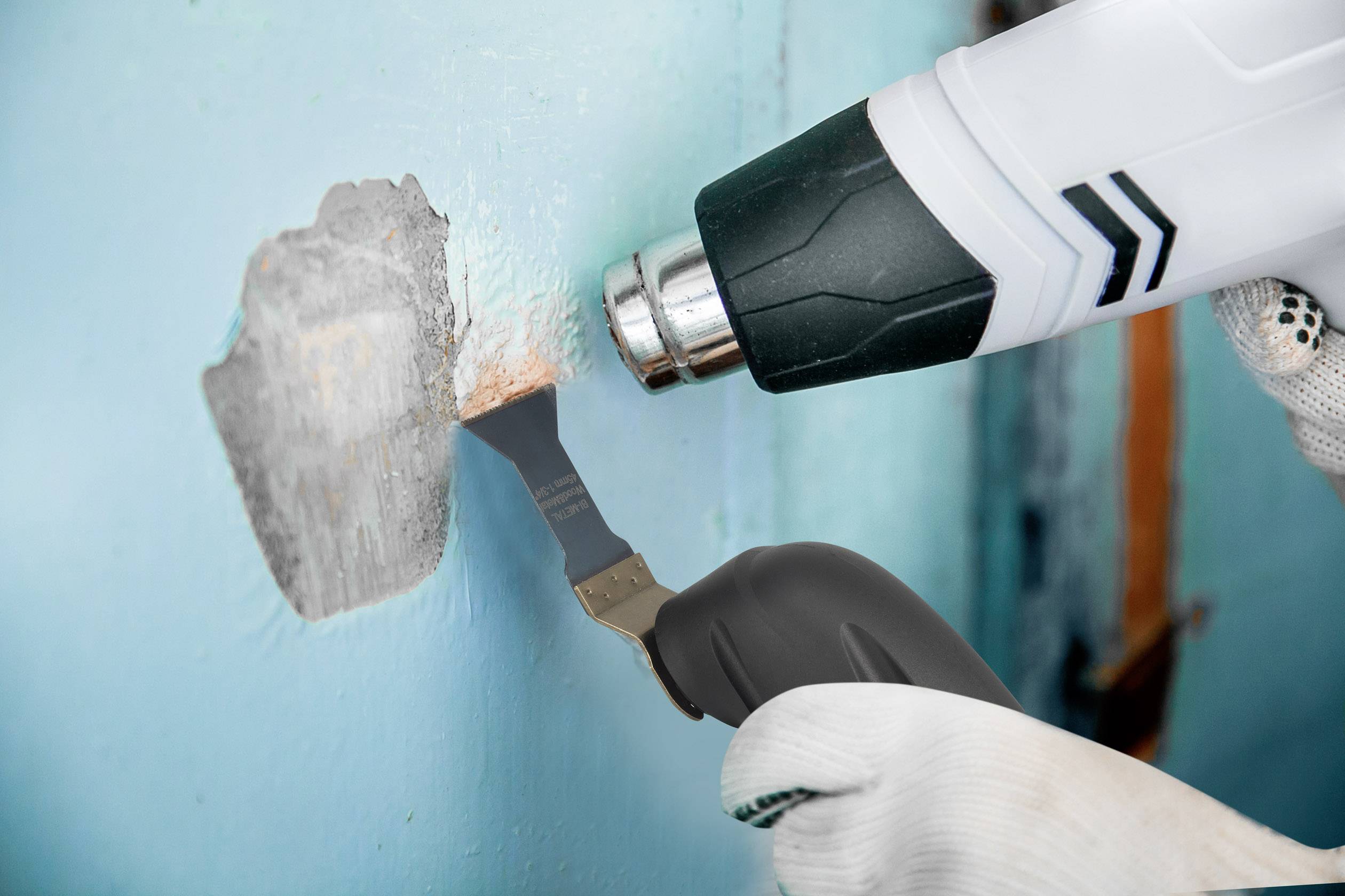 Eine Person verwendet einen Heißluftföhn und einen Spachtel, um alte Farbe von einer blauen Wand zu entfernen. Handschuhe schützen die Hände.