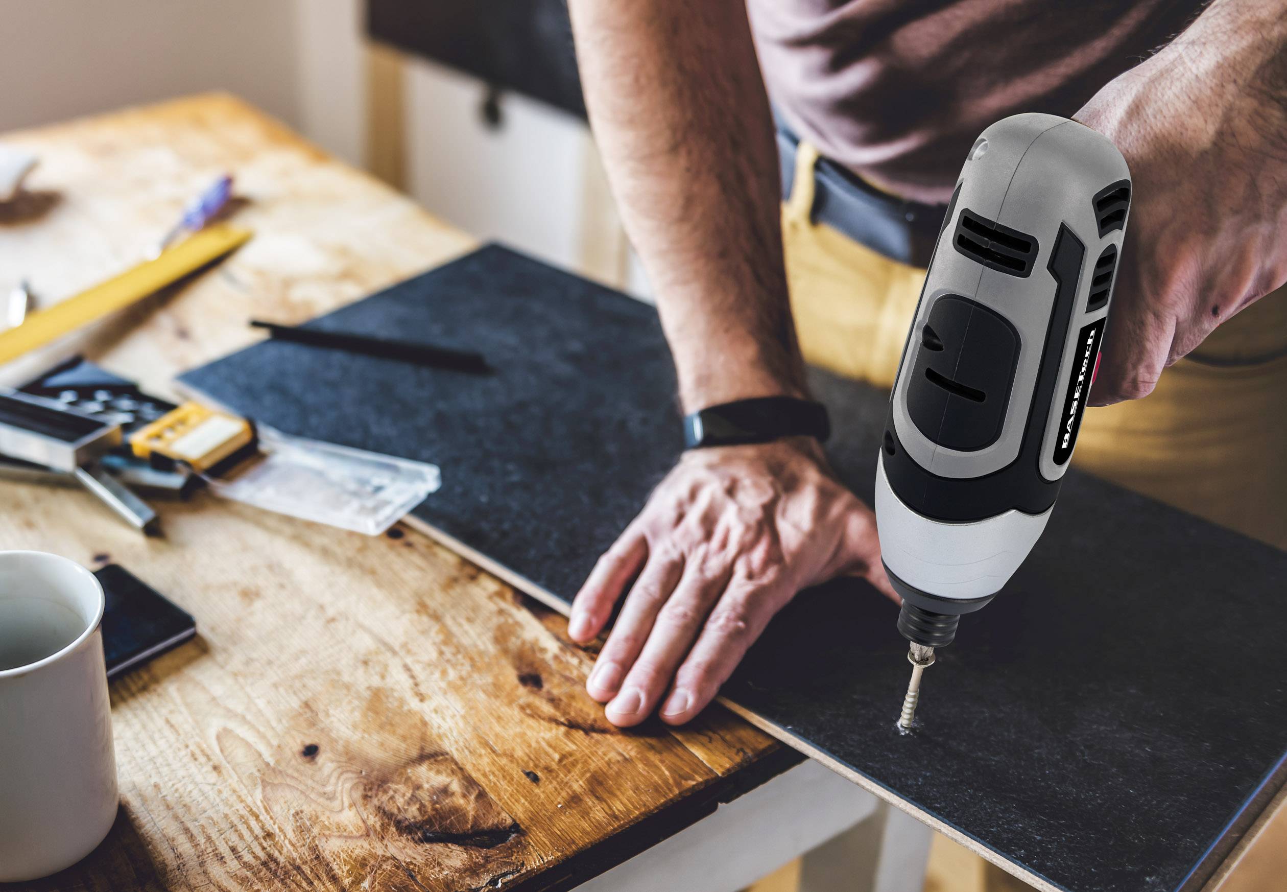Person verwendet eine elektrische Bohrmaschine, um ein Loch in eine Holzplatte zu bohren, auf einem Tisch mit Werkzeugen.