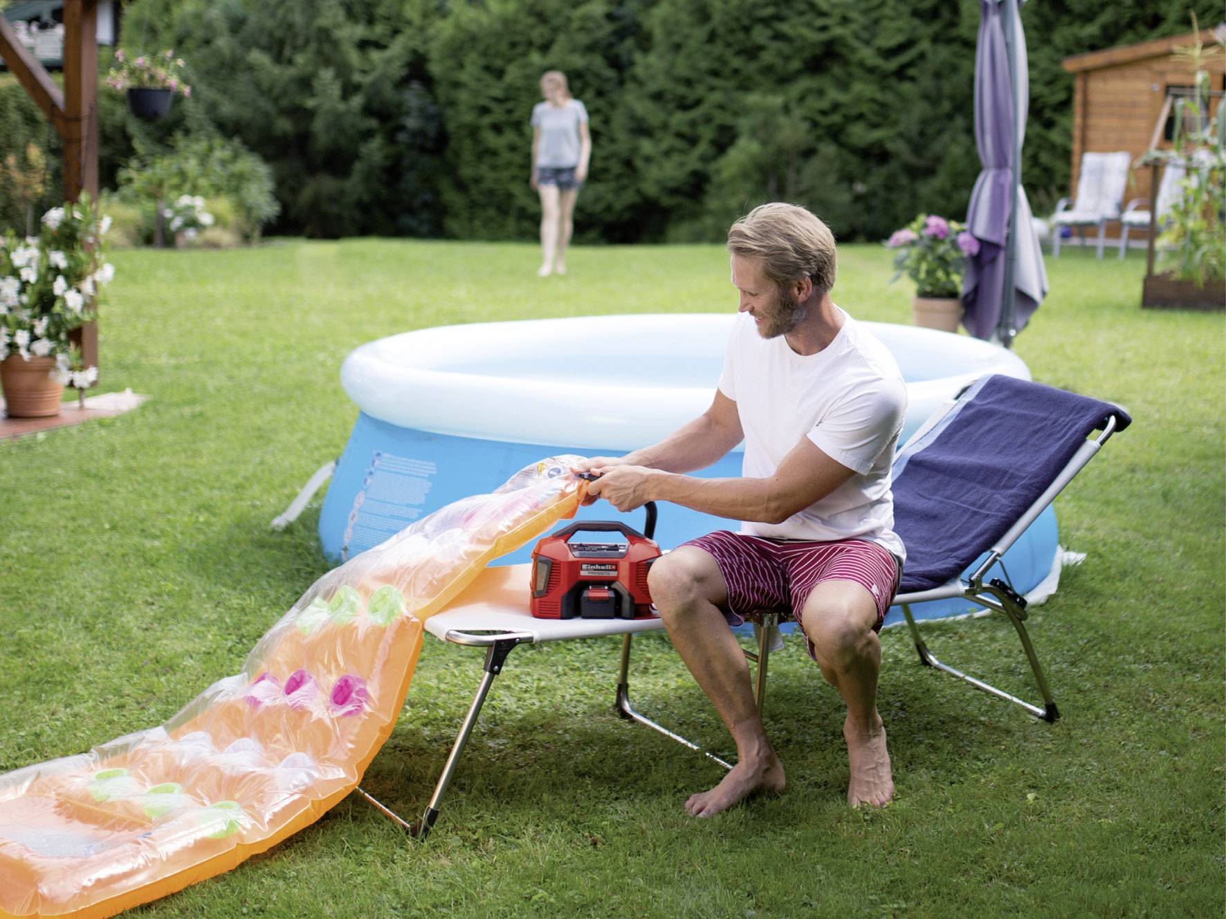 Ein Mann pumpt auf einem Liegestuhl im Garten eine Luftmatratze auf. Im Hintergrund steht ein aufblasbarer Pool.