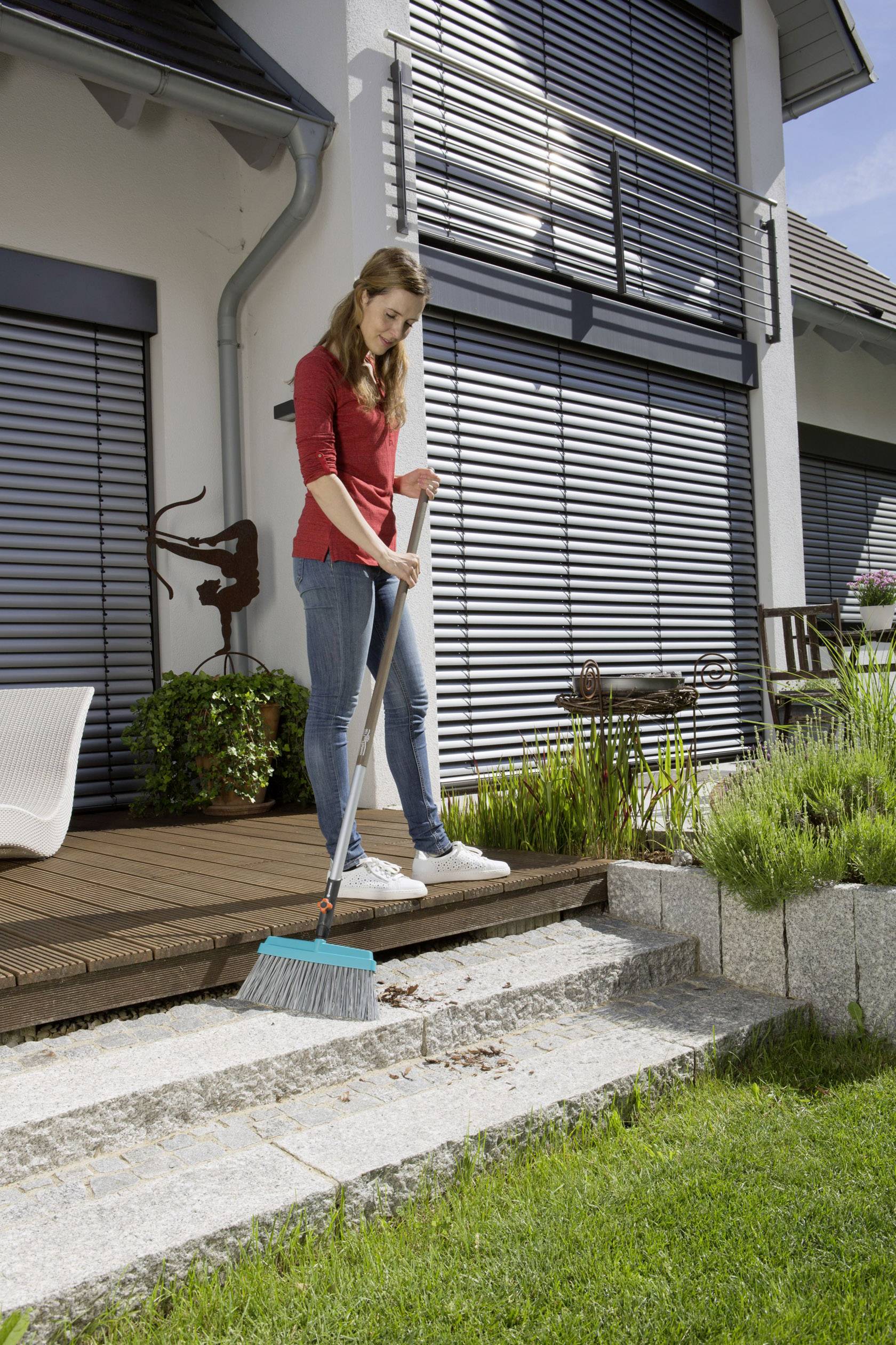 Eine Frau fegt Blätter von einer Steintreppe in einem modernen Garten mit Terrasse. Im Hintergrund ist ein Haus mit geschlossenen Jalousien.
