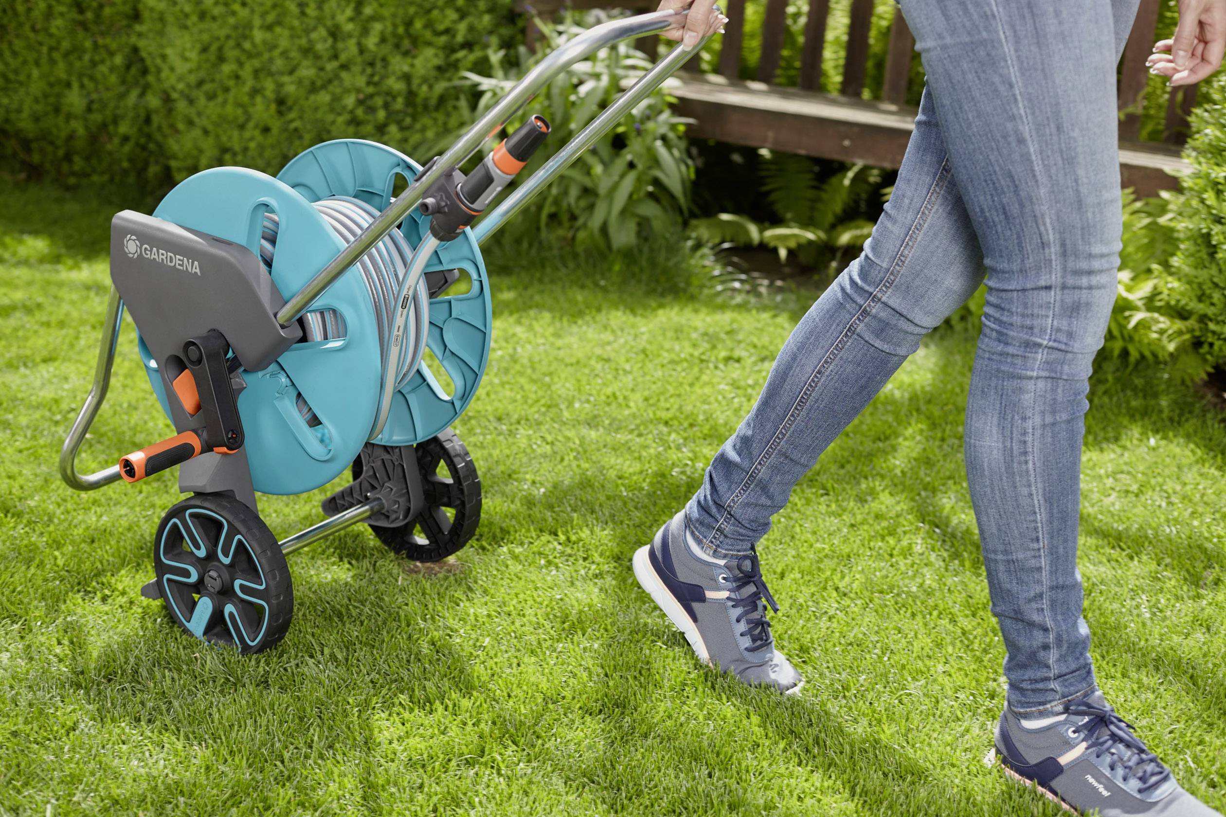 Eine Person zieht einen Gartenschlauchwagen über den Rasen. Der Schlauchwagen ist blau und hat zwei Räder zur einfachen Bewegung.