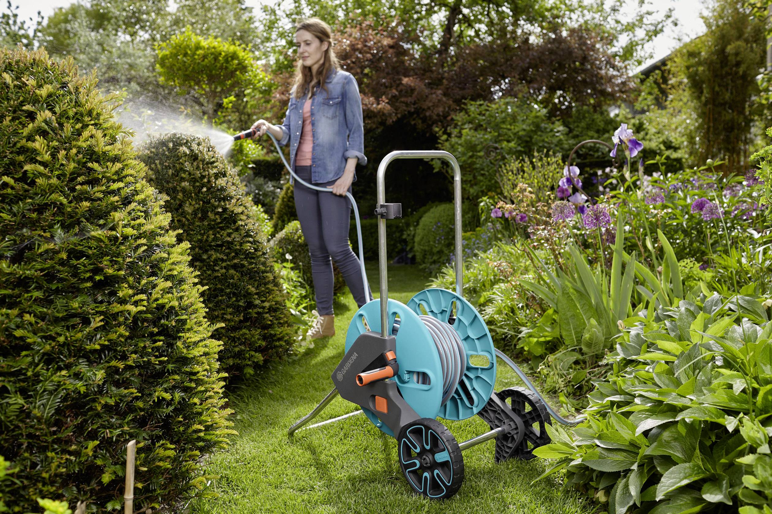Eine Frau gießt Pflanzen mit einem Gartenschlauch in einem grünen Garten. Im Hintergrund sind Büsche und blühende Blumen zu sehen.