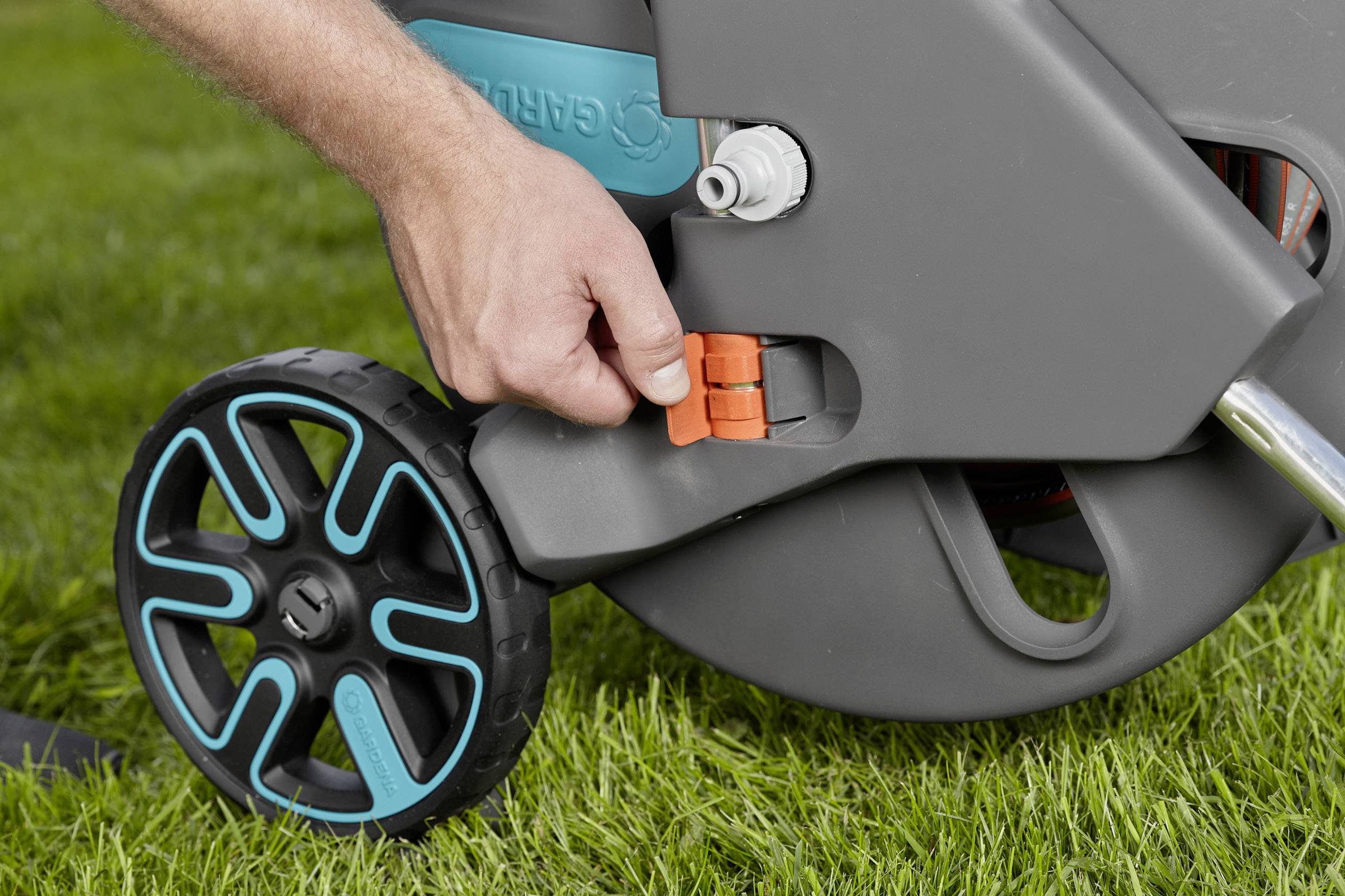 Eine Hand hält einen orangenen Hebel an einem Gartenschlauchwagen auf einer Rasenfläche. Der Schlauchwagen hat schwarze Räder mit blauen Akzenten.