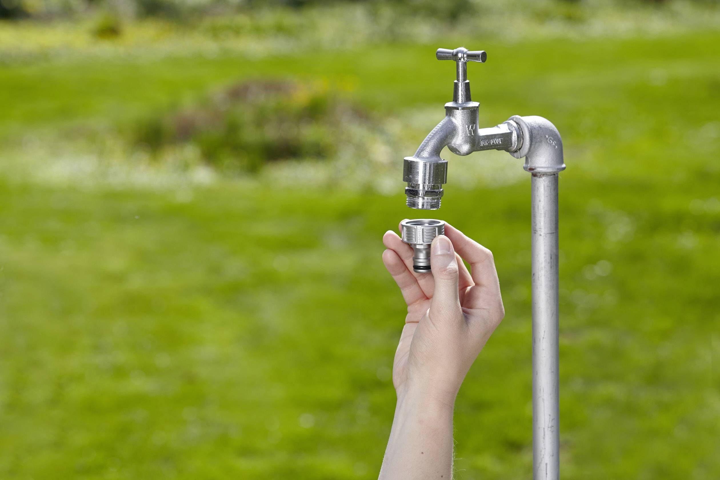 Eine Hand hält einen Metalladapter vor einen Wasserhahn im Freien, über grünem Rasen.