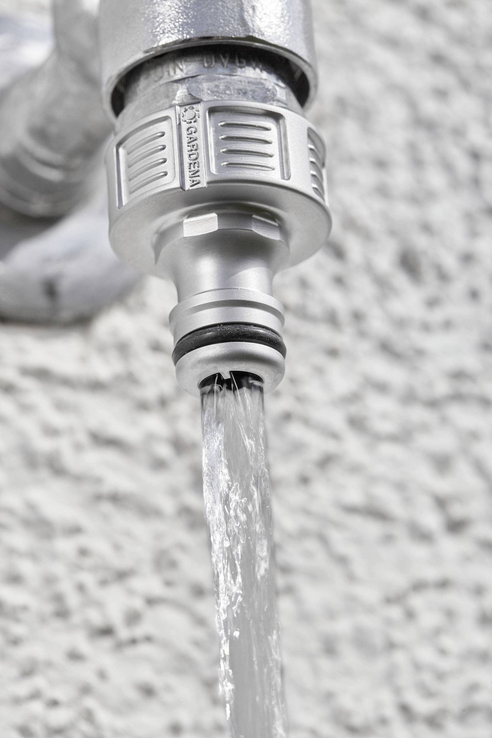Wasser fließt aus einem silbernen Wasserhahn an einer grauen Wand. Der Fokus liegt auf dem Wasserstrahl und der Hahnverbindung.