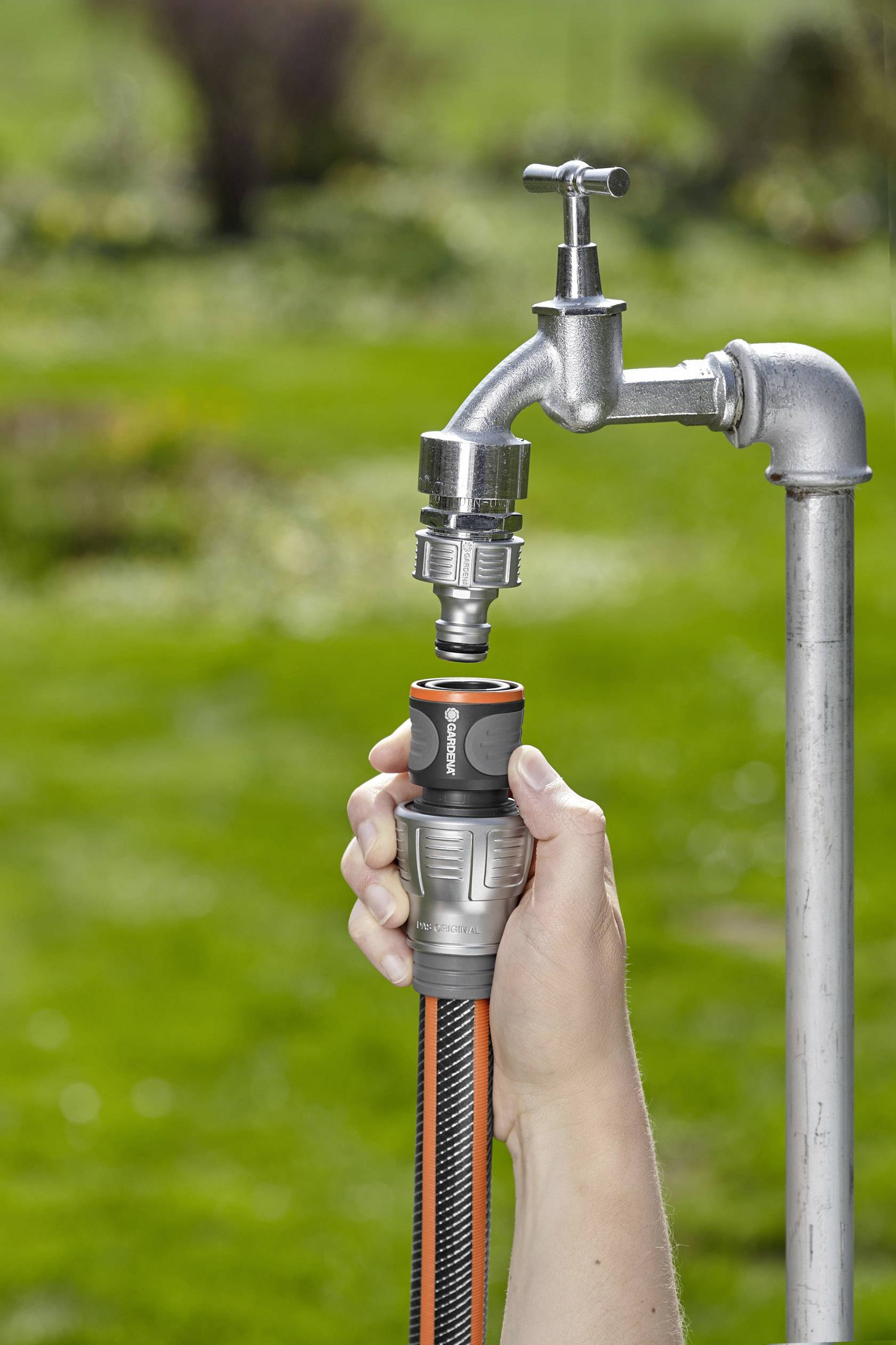 Hand verbindet Gartenschlauch mit Wasserhahn im Garten. Im Hintergrund ist unscharfes Grün zu sehen.