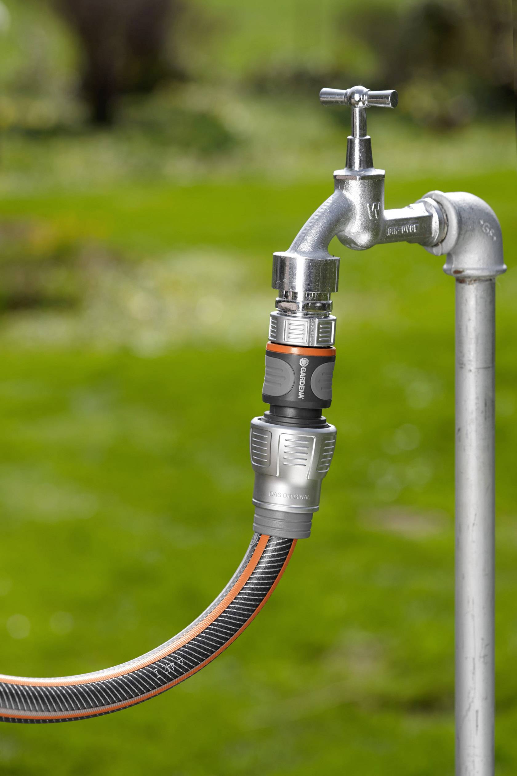 Ein silberner Wasserhahn im Freien mit daran angeschlossenem Gartenschlauch mit orange-grauer Kupplung, vor grünem Hintergrund.