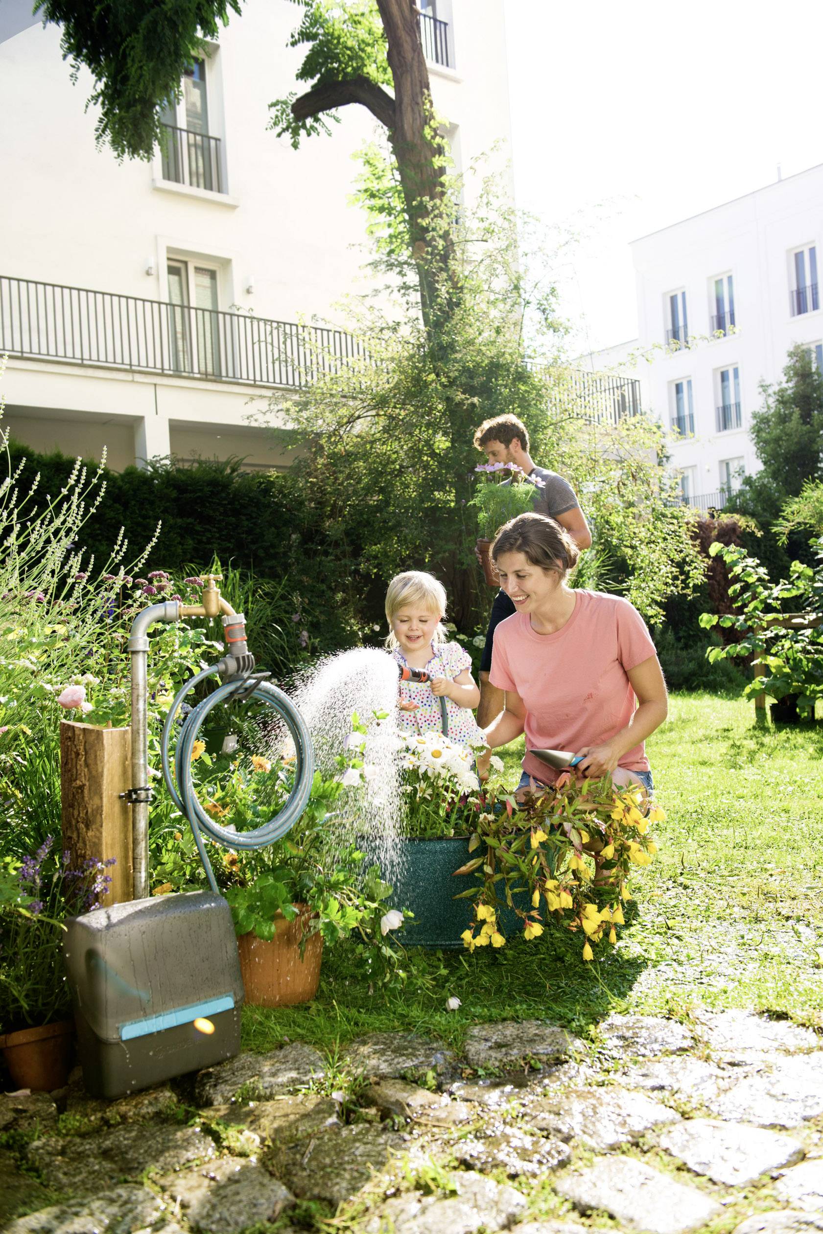 Eine Familie bewässert den Garten: Eine Frau gießt Blumen, ein Kind hält den Gartenschlauch, während ein Mann im Hintergrund steht.