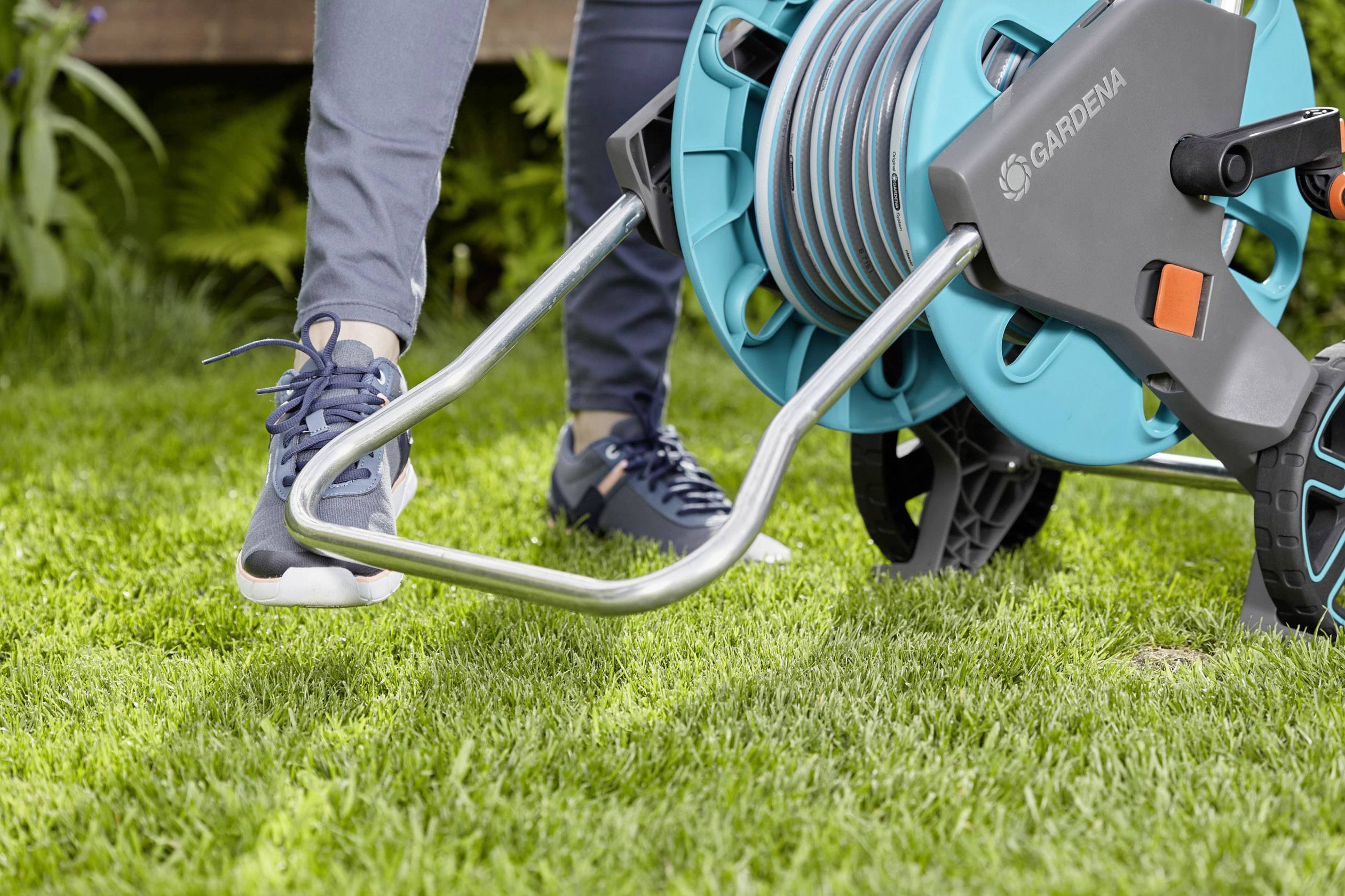 Eine Person benutzt eine Gartenschlauchrolle auf einem Rasen. Die Person trägt sportliche Kleidung und bewegt den Schlauchwagen.