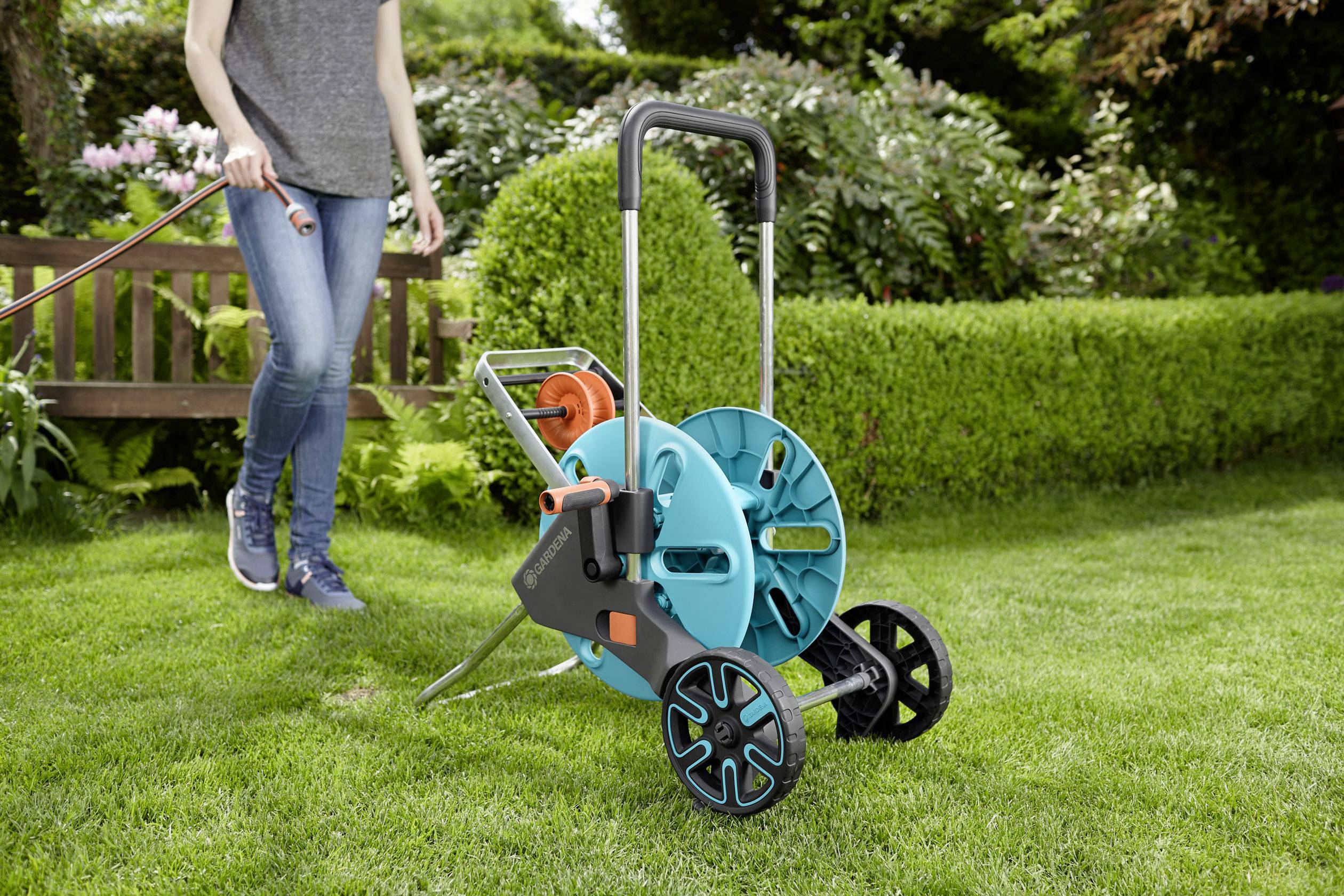Eine Person im Garten zieht einen Gartenschlauchwagen über den Rasen. Im Hintergrund sind grüne Büsche und eine Holzbank zu sehen.