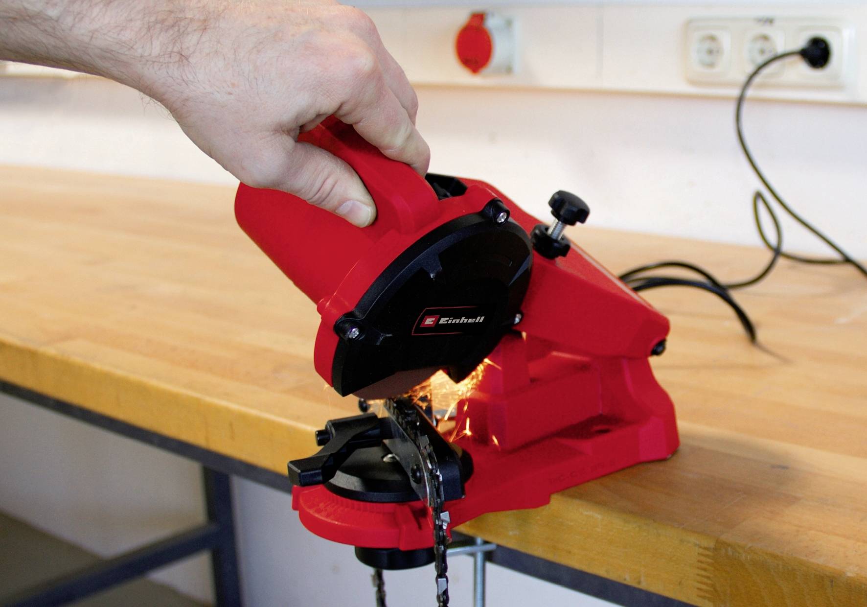 Eine Hand bedient einen roten Kettenschärfer auf einer Werkbank. Funken sprühen, während die Kette geschärft wird.